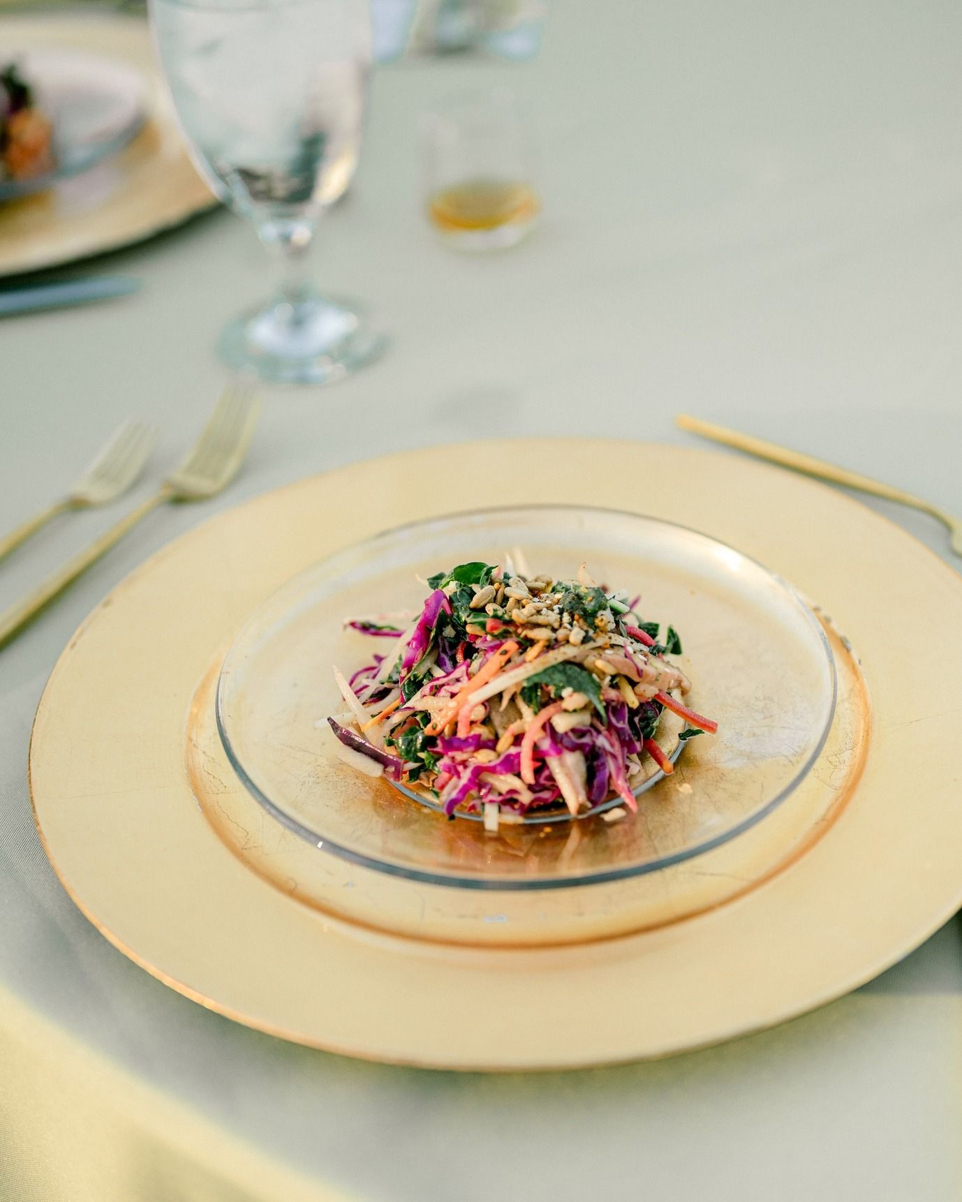 Colorful slaw salad on a small glass plate, atop a gold charger. Includes forks, a glass of water, and a gold tablecloth.