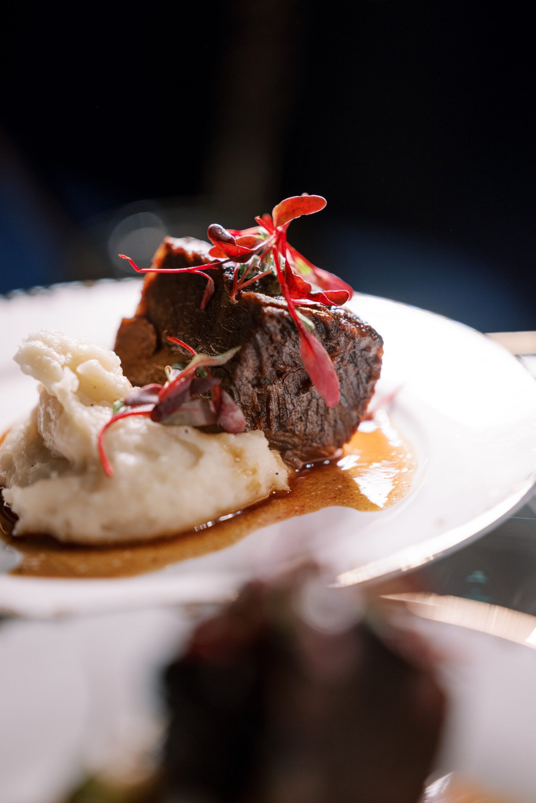 Braised beef on mashed potatoes, garnished with red petals, served on a white plate with a dark background.