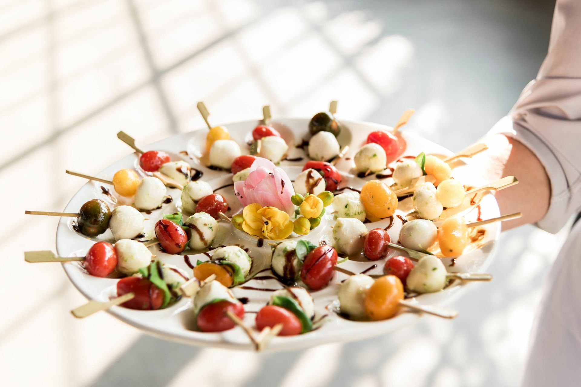 Person holding a white plate with small skewers of mozzarella balls, cherry tomatoes, and basil drizzled with balsamic glaze.