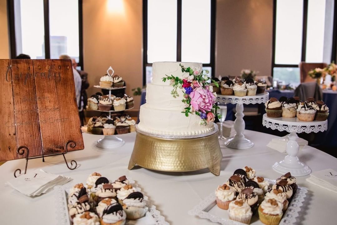 A wedding cake table with a three-tiered cake decorated with flowers, cupcakes, and pastries, set in a venue with windows.