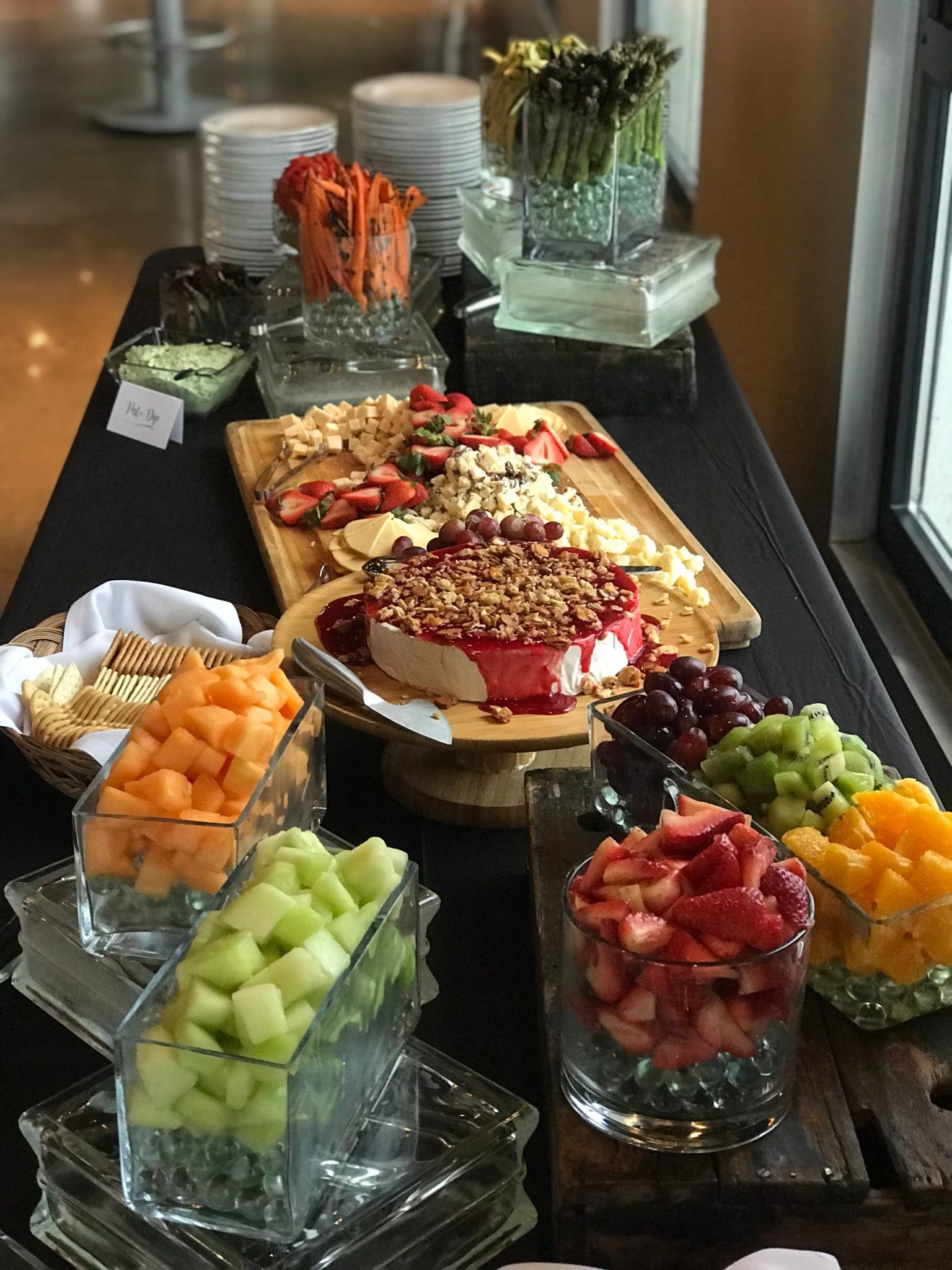 A buffet table with various foods like fruit, cheese, and vegetables arranged for a party or event. There is a dark background with light shining in from the side.