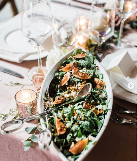 Oval serving dish filled with green salad and orange fruit on a pink tablecloth, near candles and glassware, set for dining.