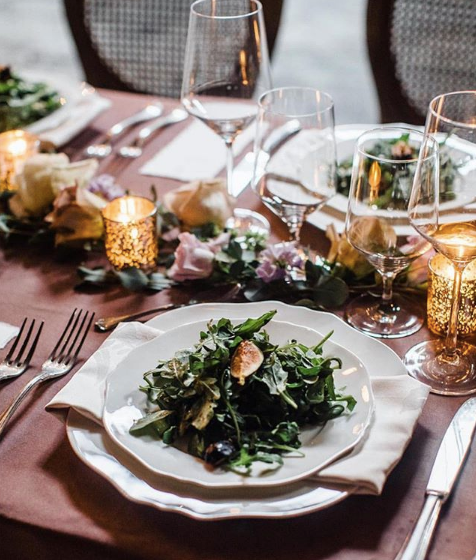 Elegant table setting with salad plates, wine glasses, flowers, and candles. Dark tablecloth.