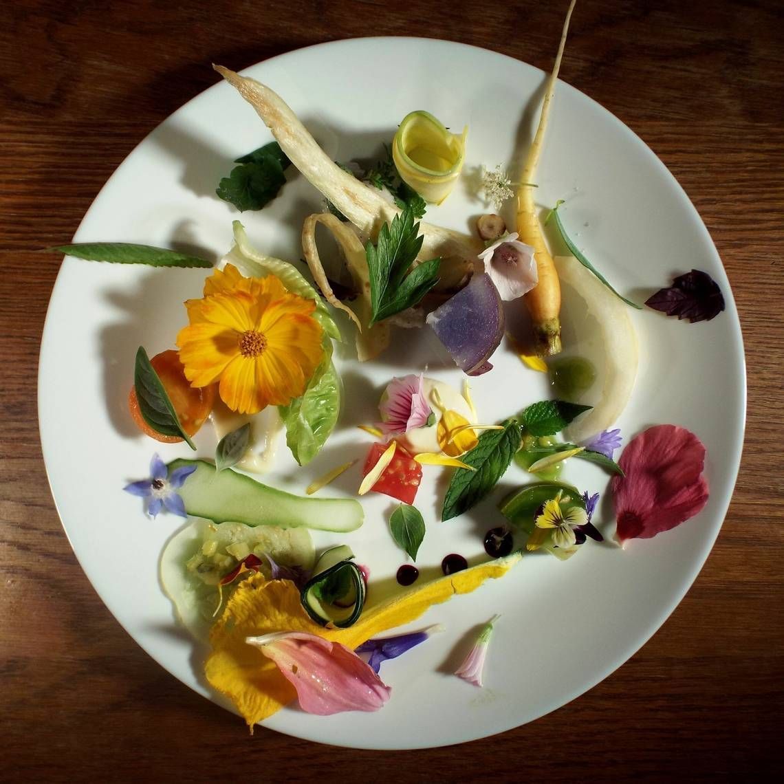 White plate with a colorful arrangement of edible flowers, herbs, and sliced vegetables, on a wooden table.