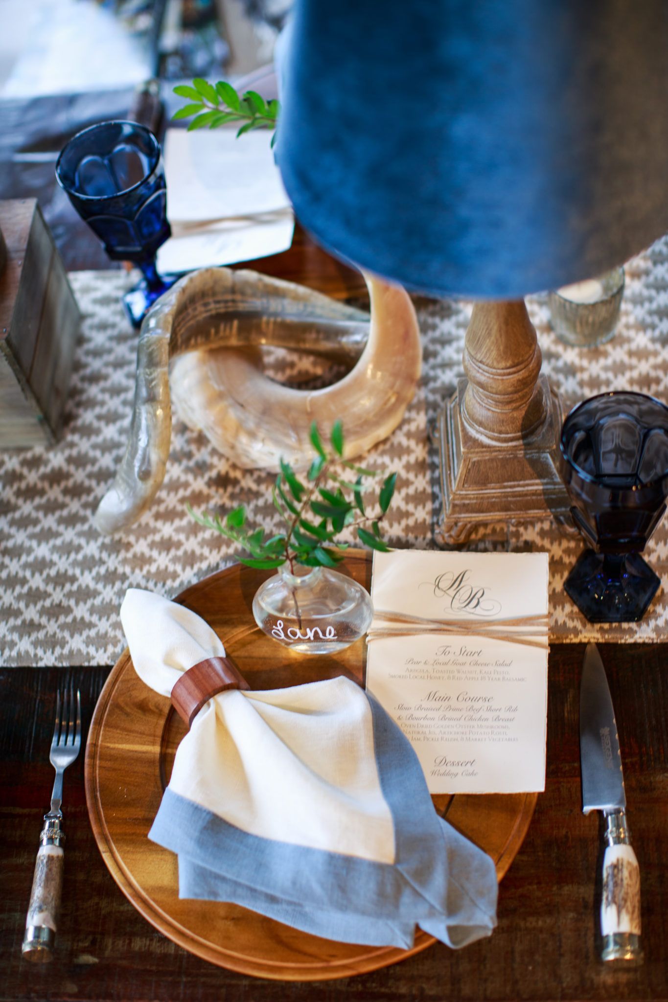 A table setting with a wooden plate, blue-trimmed napkin, and menu. Includes silverware, a ram's horn, and blue glassware.