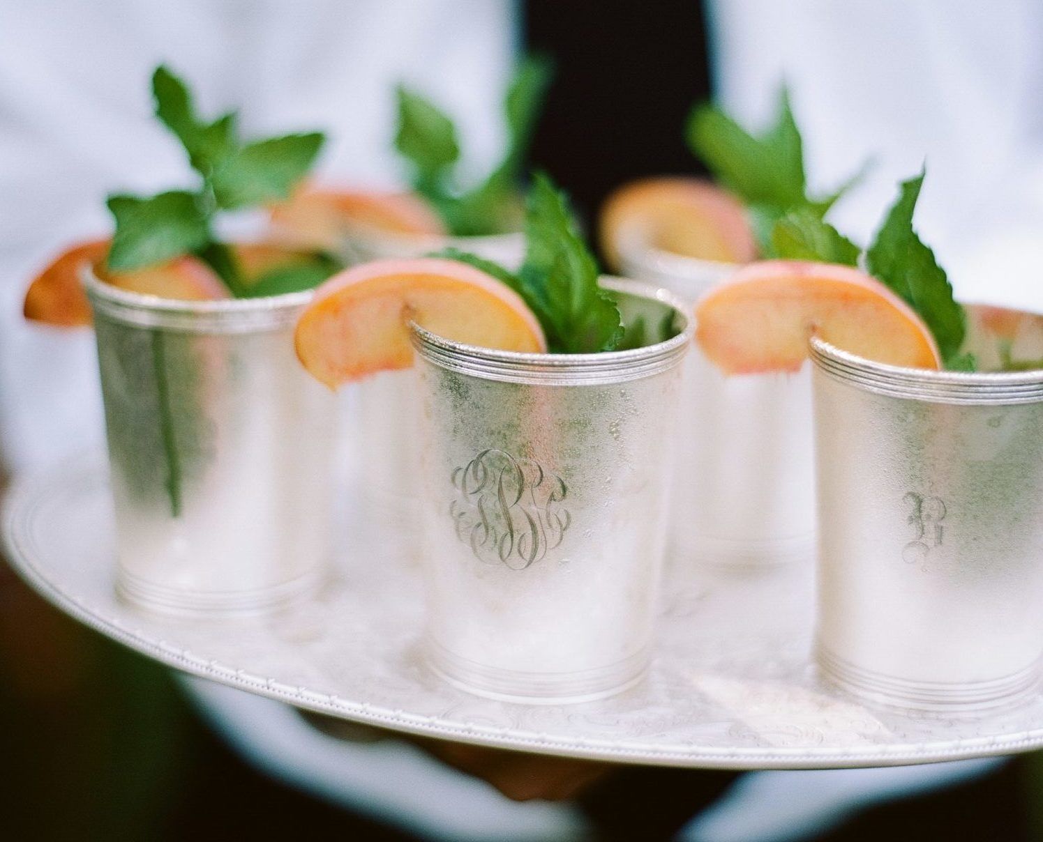 A server holding a tray of silver cups garnished with peach slices and mint sprigs.