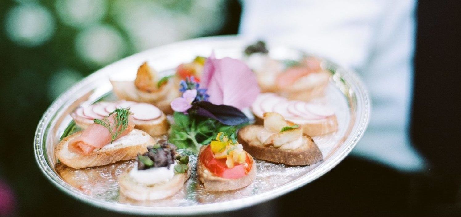 A platter of appetizers, with various toppings and garnishes, is held by a person with a blurred green background.