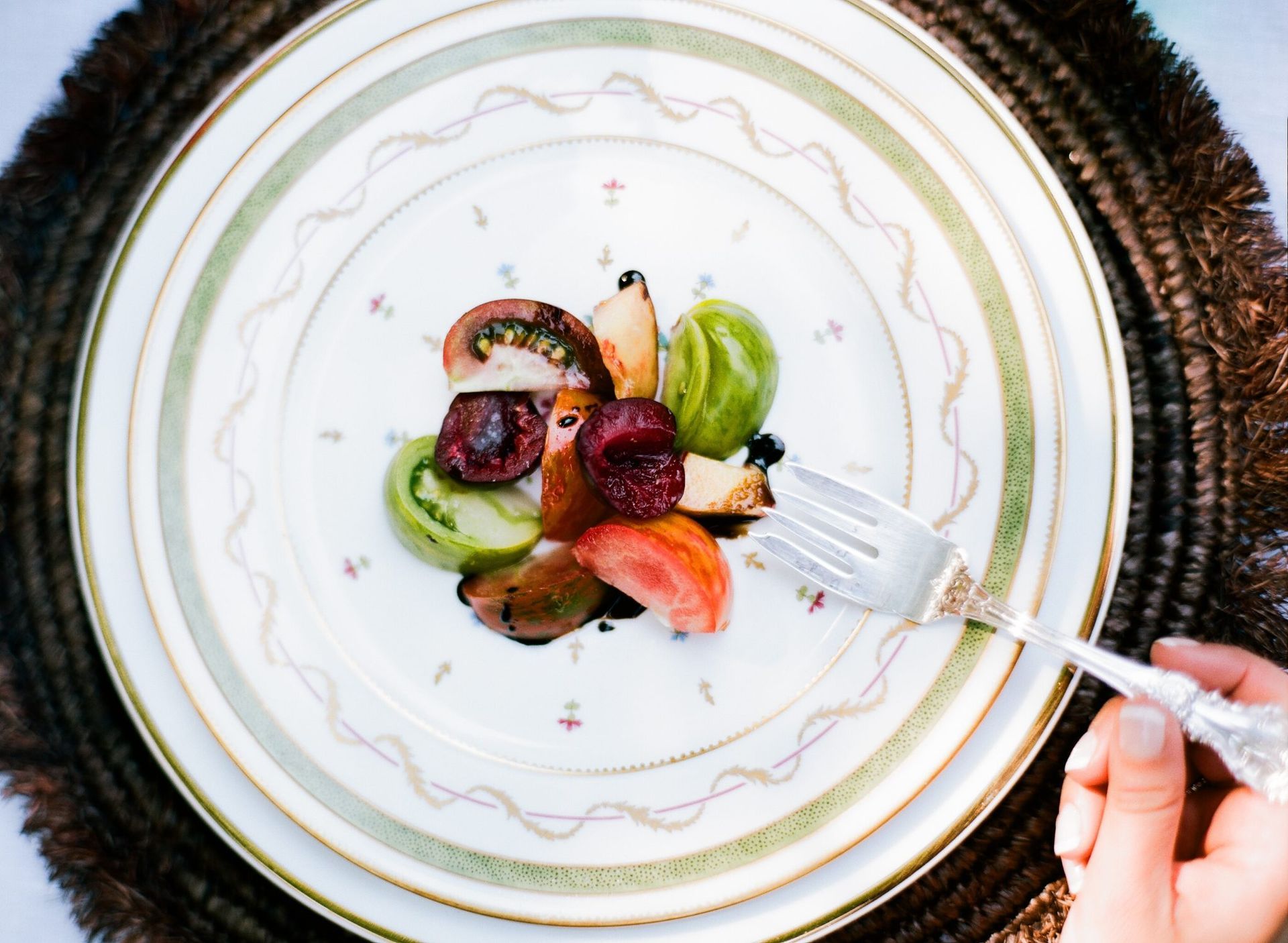 Sliced heirloom tomatoes on a decorative plate with a silver fork and a person's hand, set on a woven mat.