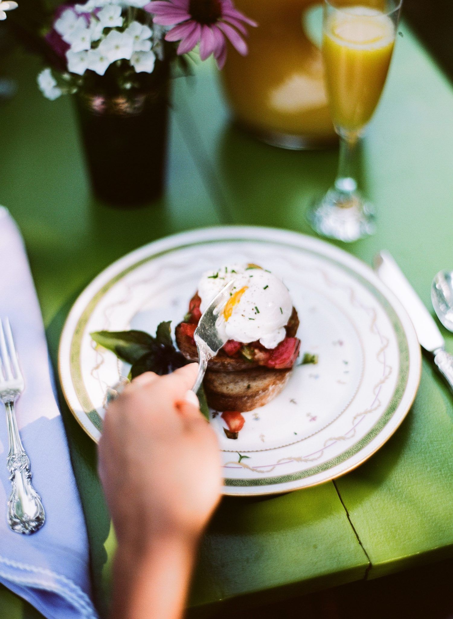 Breakfast plate with a poached egg, hand reaching for it on a green table. Mimosa and flowers in background.