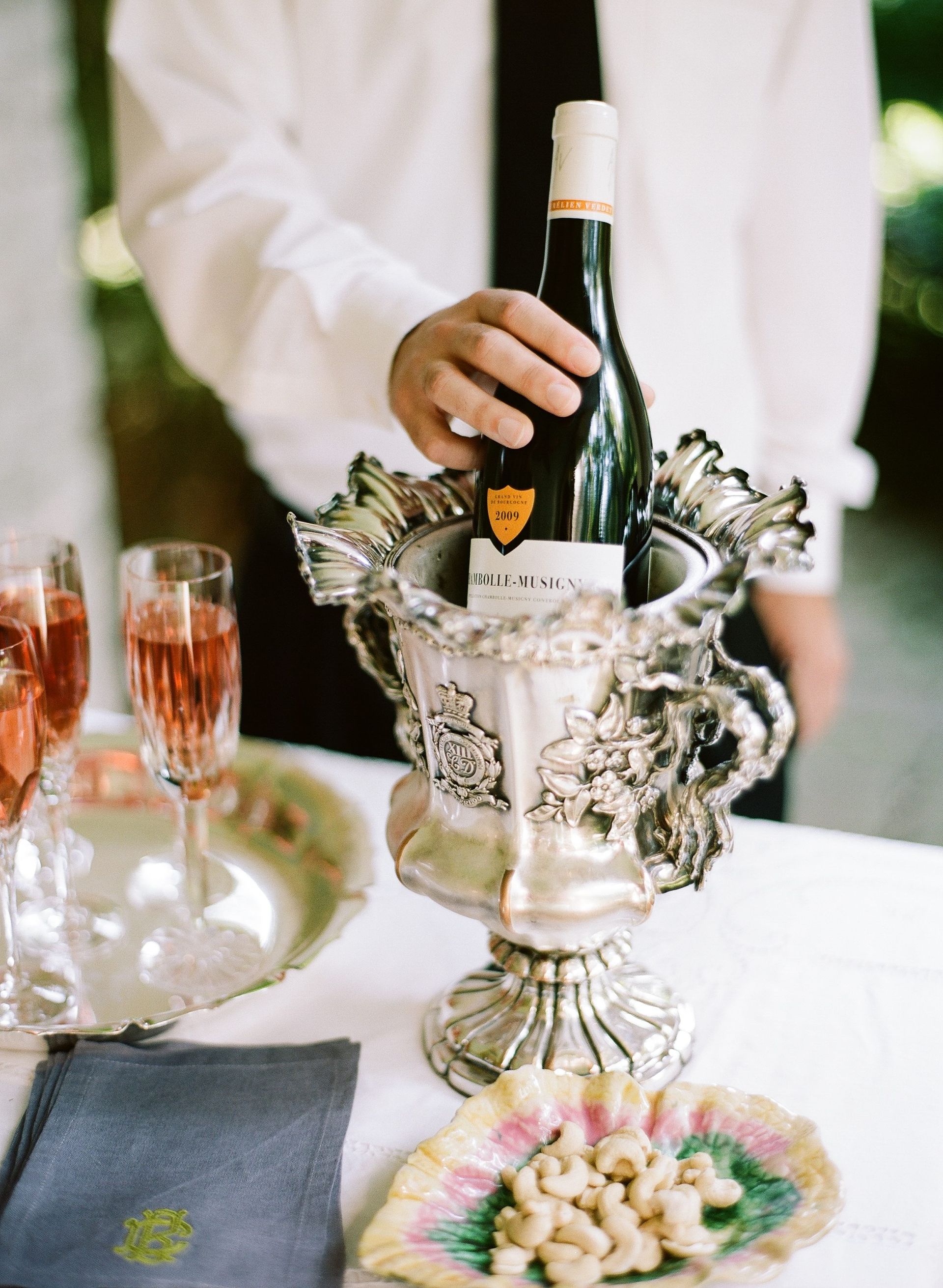 Person in white shirt and tie holding a wine bottle in an ornate silver ice bucket, champagne flutes to the left.