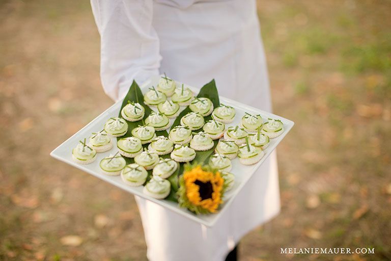 A server holds a tray of appetizers, each topped with a green filling and garnish, a sunflower adding color to the arrangement.