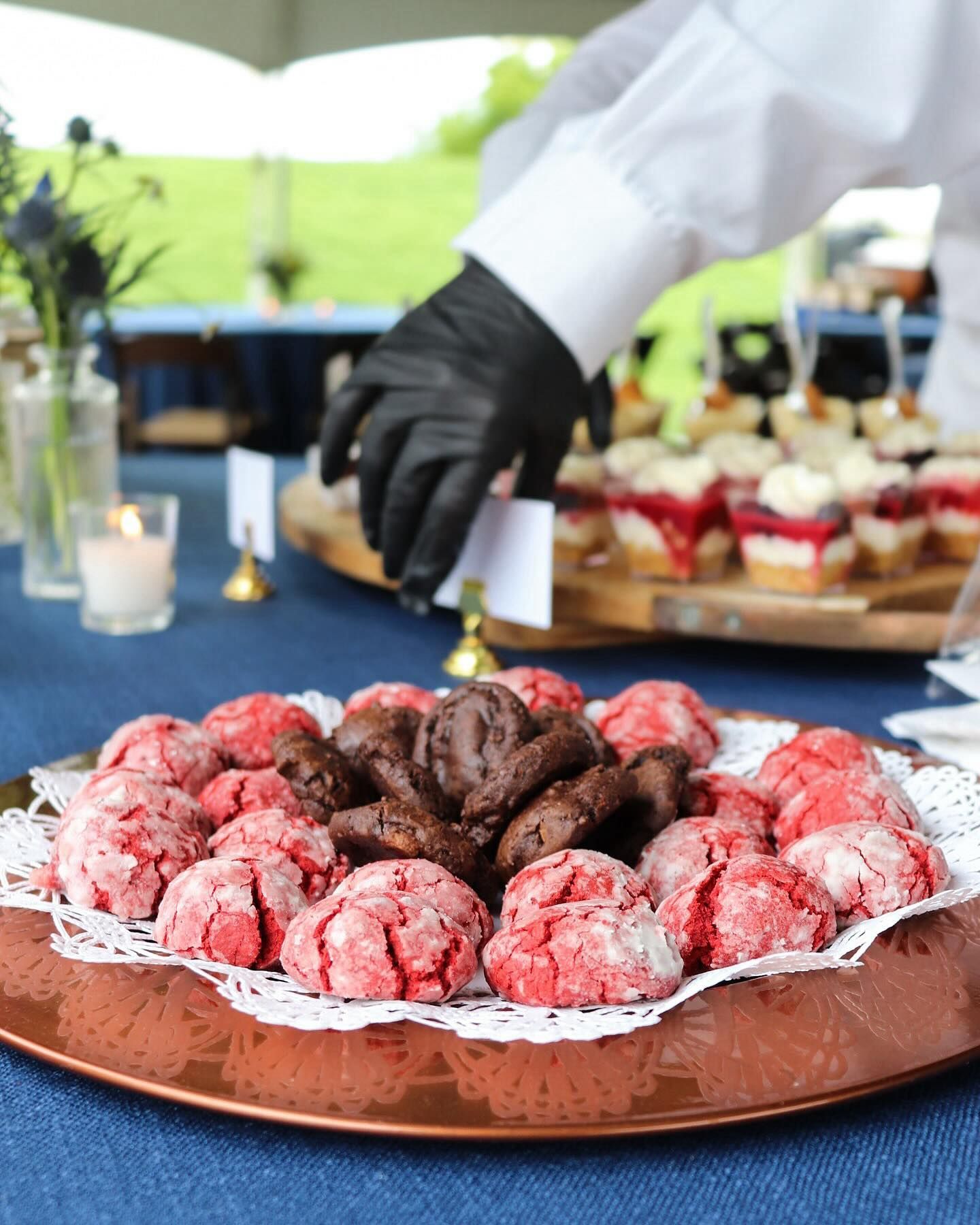 Tray of red and brown cookies on a table with desserts and a person in a white shirt and black glove reaching for them.