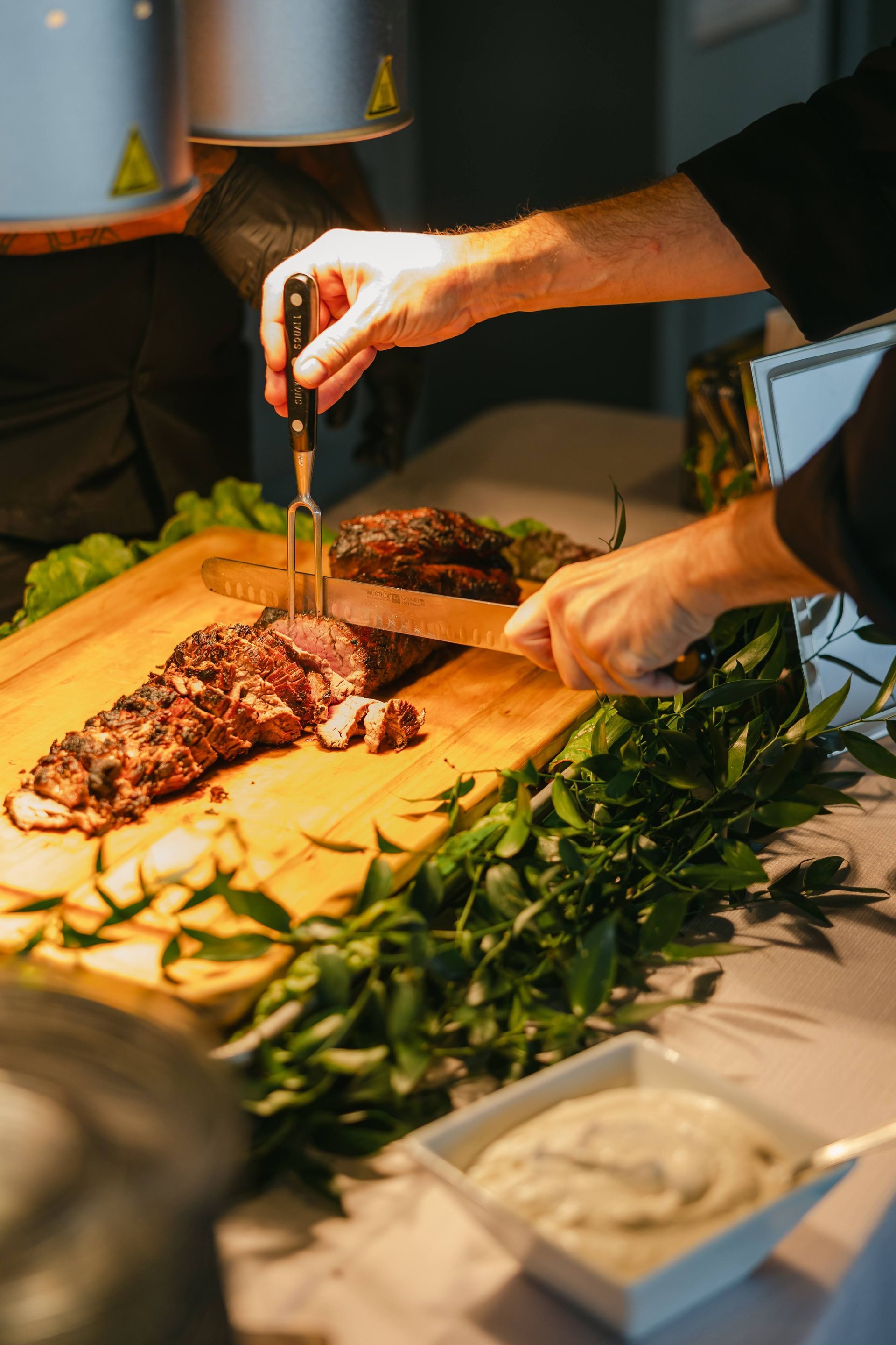 Chef carving roasted meat on a wooden board garnished with greenery, alongside a serving dish of sauce.