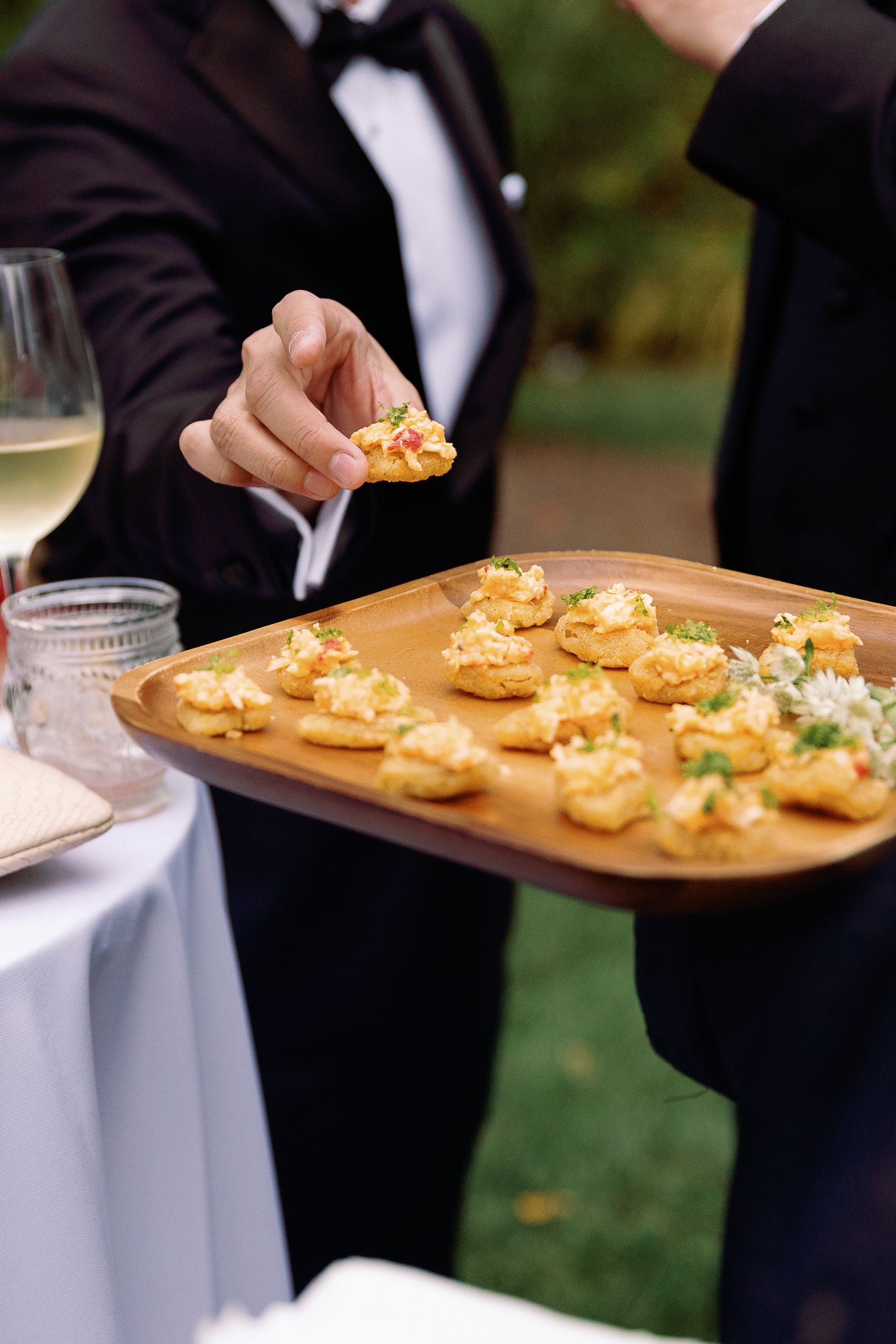 A person in a tuxedo offers a cracker topped with food from a wooden tray at an outdoor event. Another person in a tuxedo stands nearby.
