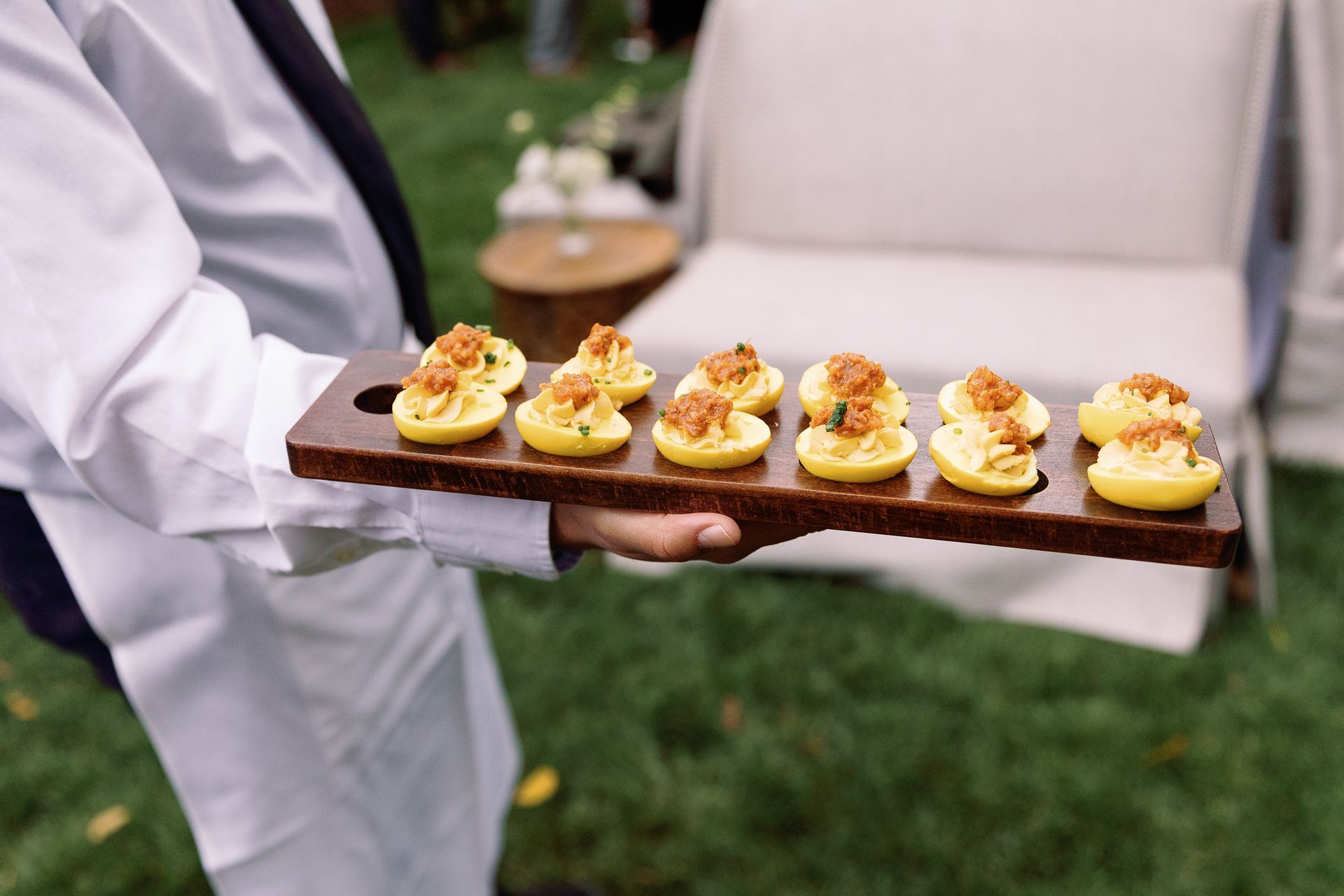 A server holds a wooden tray of deviled eggs outdoors. The eggs are yellow and topped with a reddish-brown garnish, set against a green lawn.