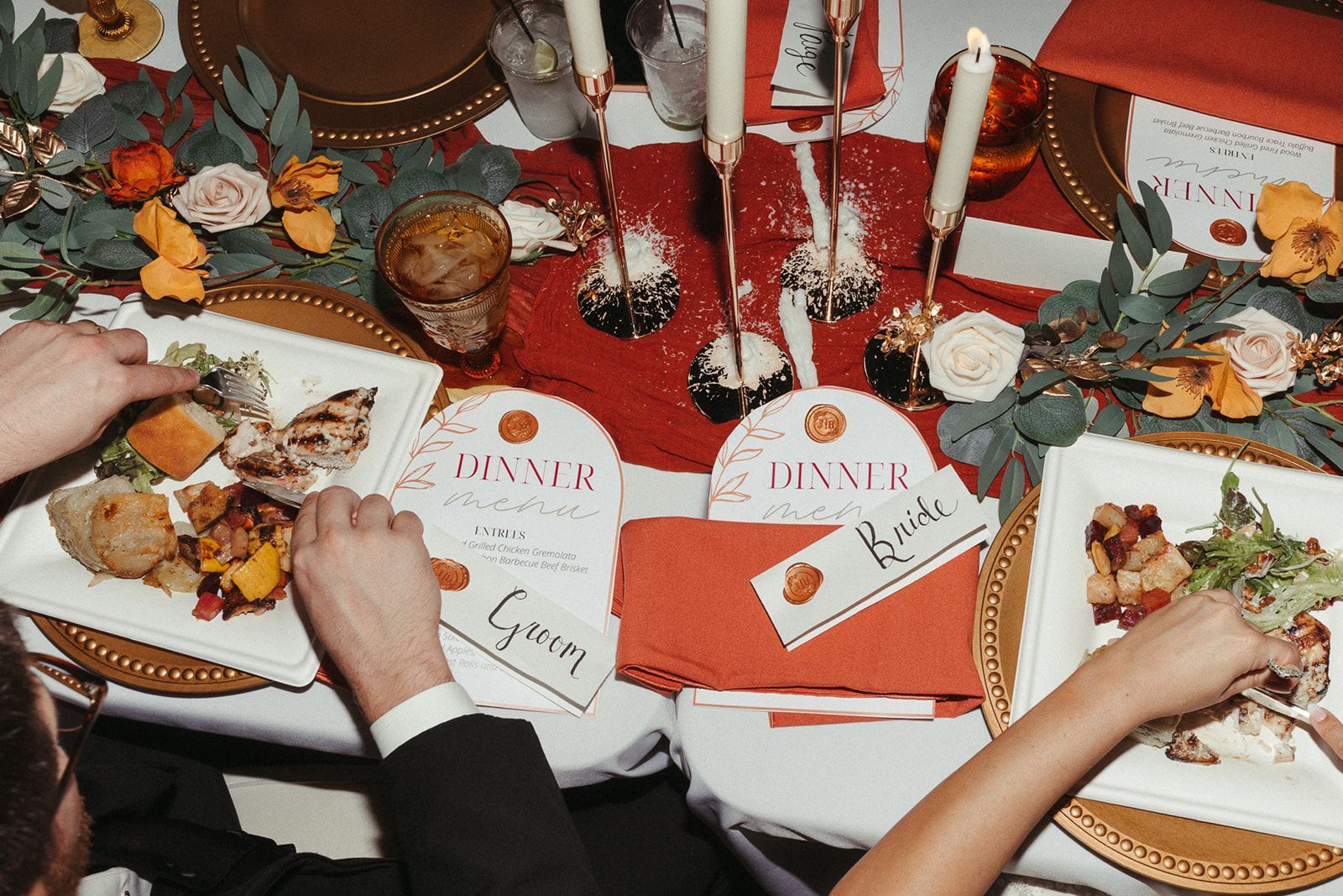 Formal dinner table setting with guests reaching for food on gold-rimmed plates. Festive decor, including flowers, candles, and personalized place cards.