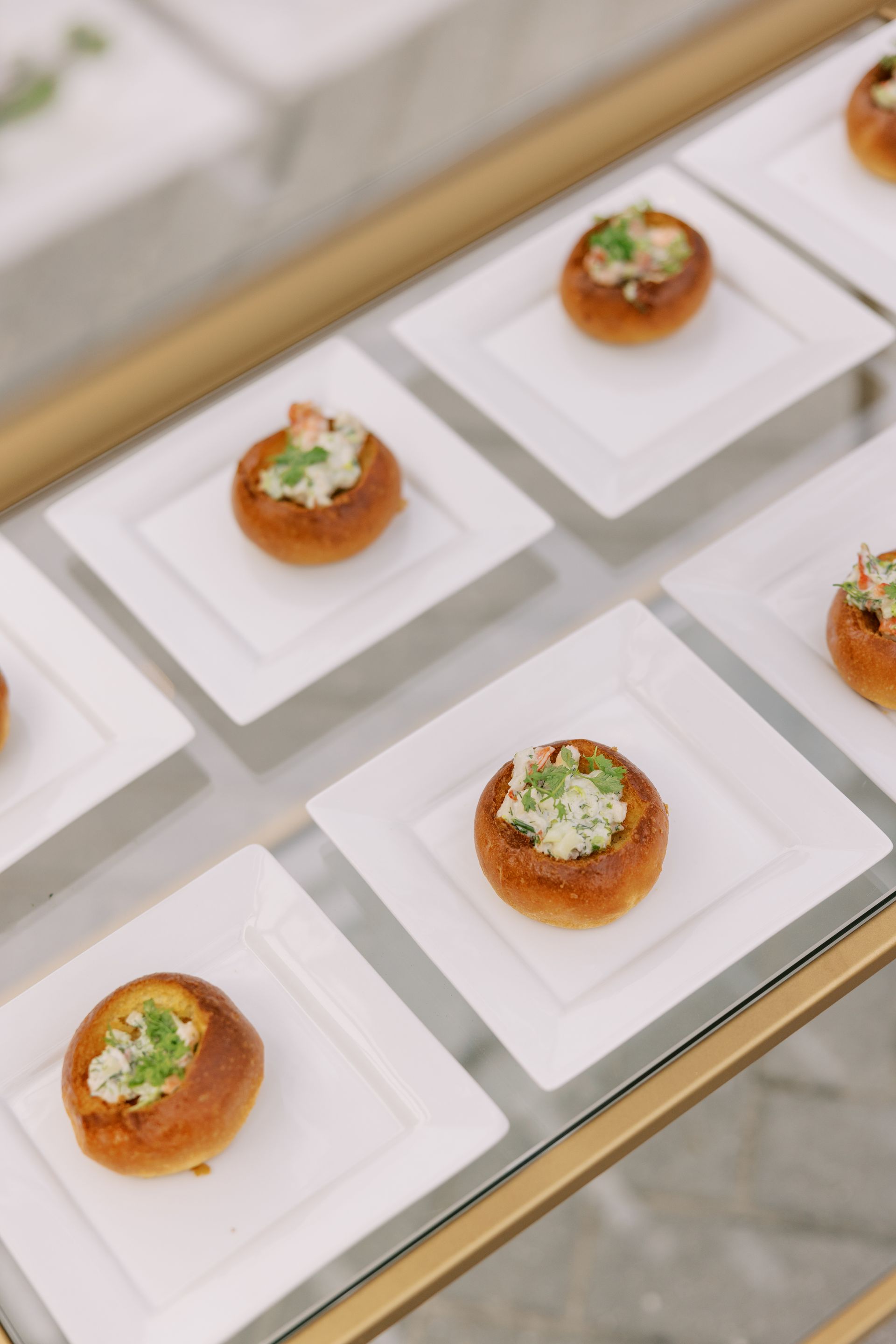 Small, bread-like appetizers topped with a creamy filling and herbs, arranged neatly on white square plates displayed on a glass surface.