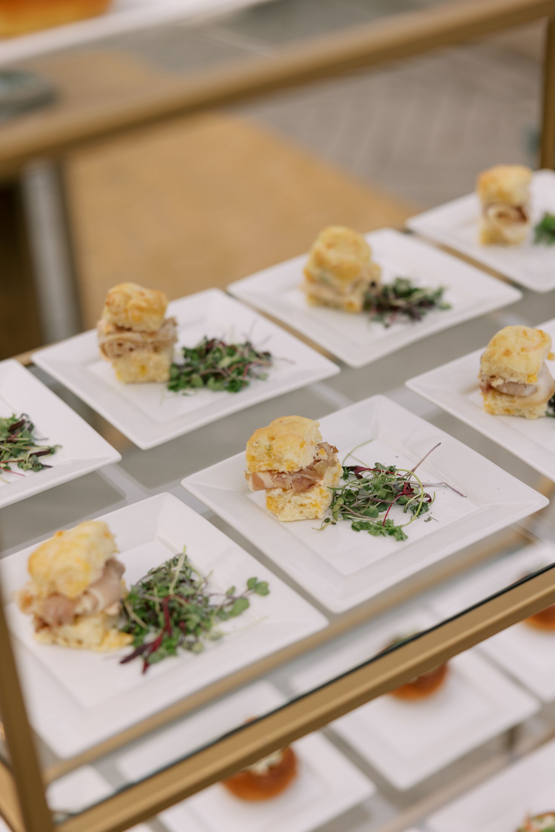 A tray of square white plates each holding a biscuit sandwich and greens. The setting is a food display at an event.