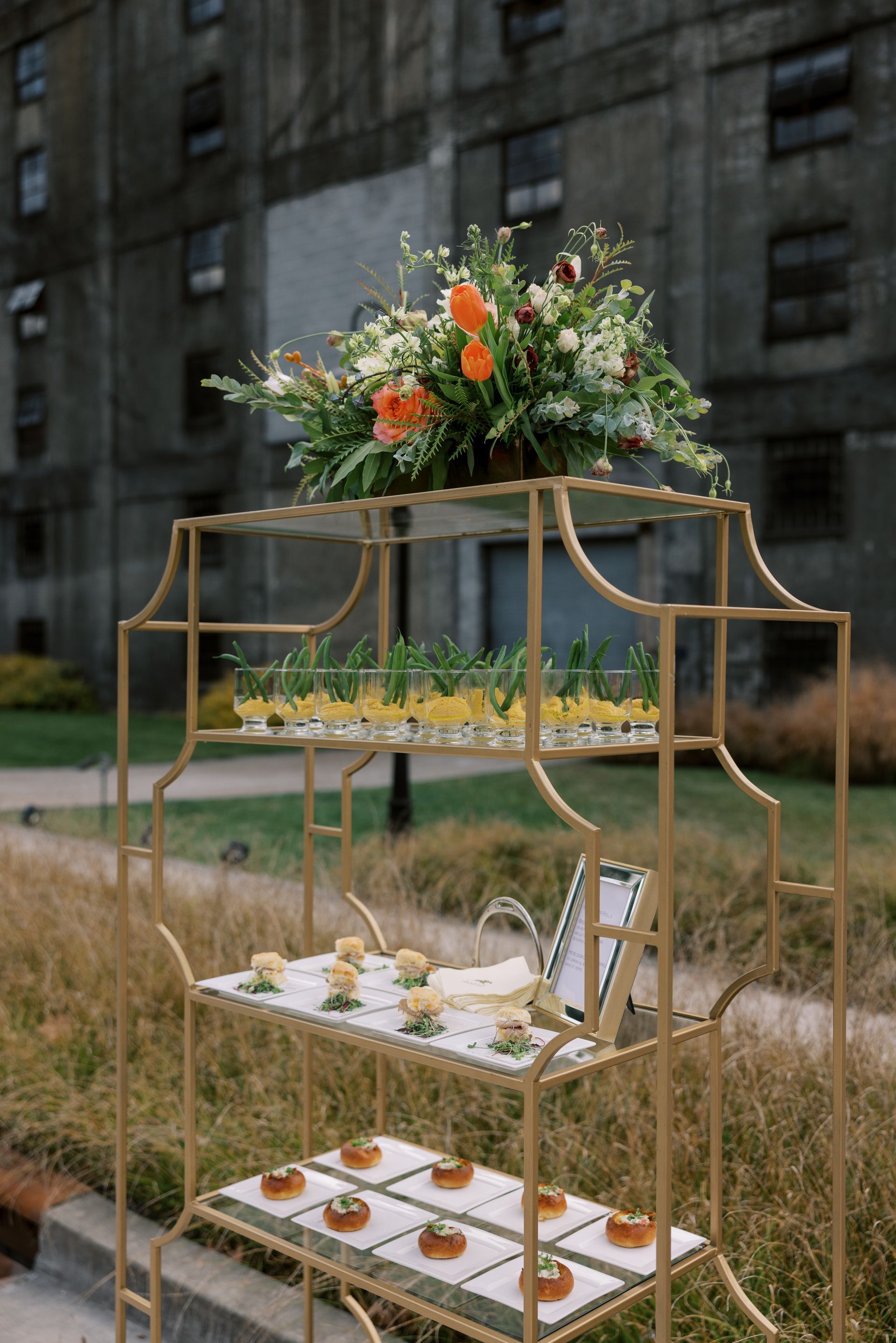A gold tiered serving cart with appetizers and a floral arrangement.  An old brick building is in the background.