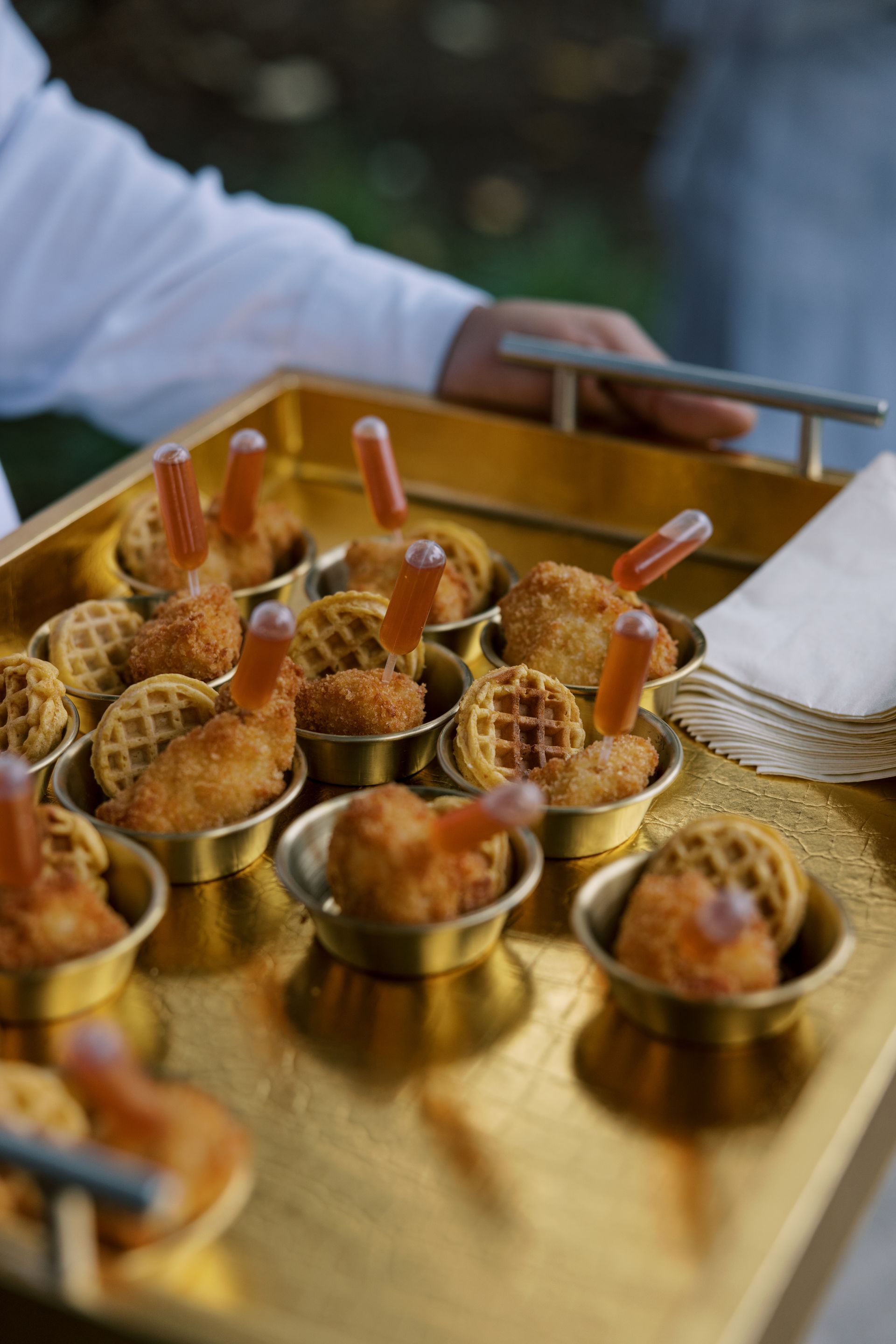 Golden tray of mini chicken and waffles, with sauce and napkins, being served.