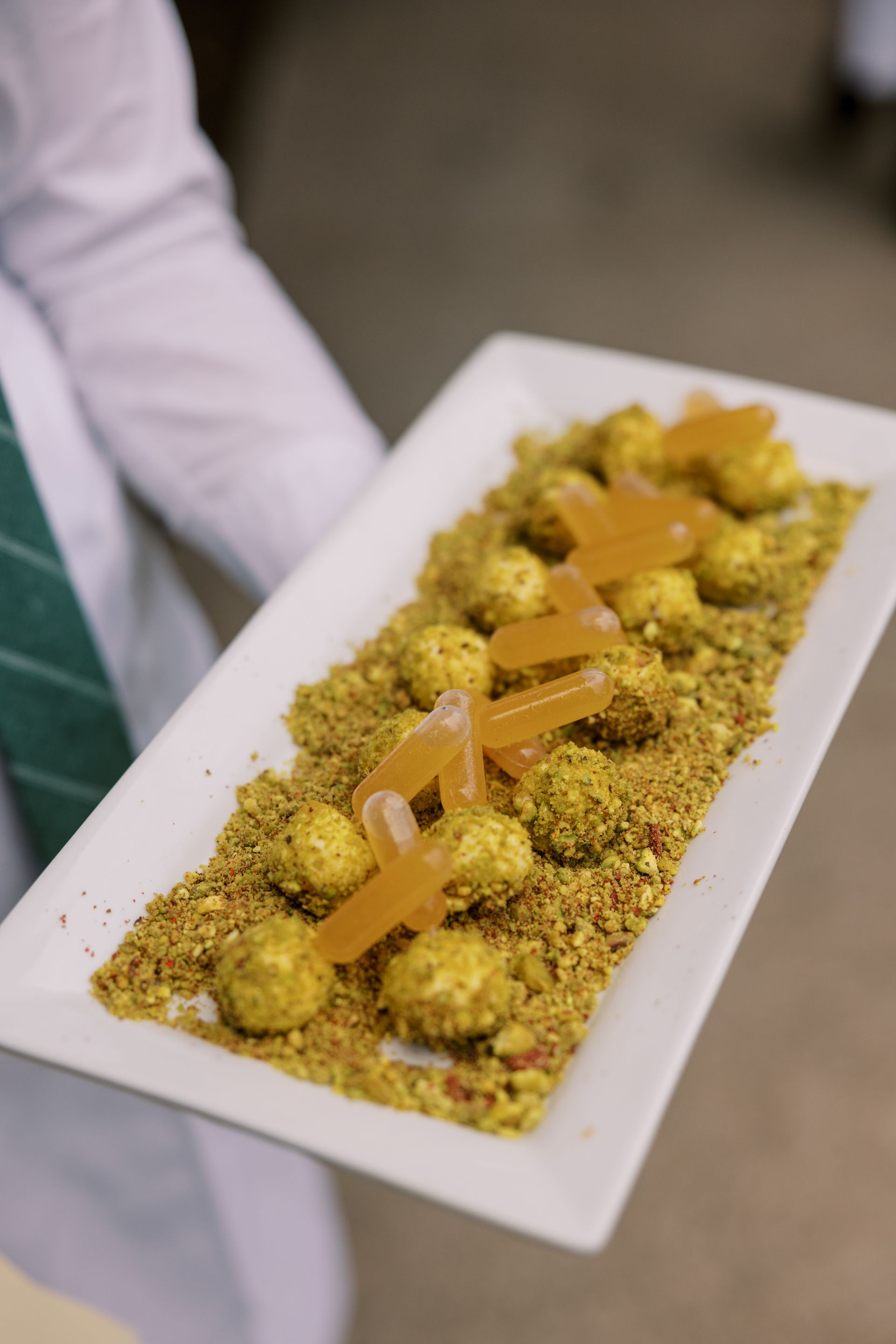 A server holds a rectangular white plate of food. The dish consists of small, round, yellow balls coated in a green-brown crumble and topped with caramel-colored pieces.