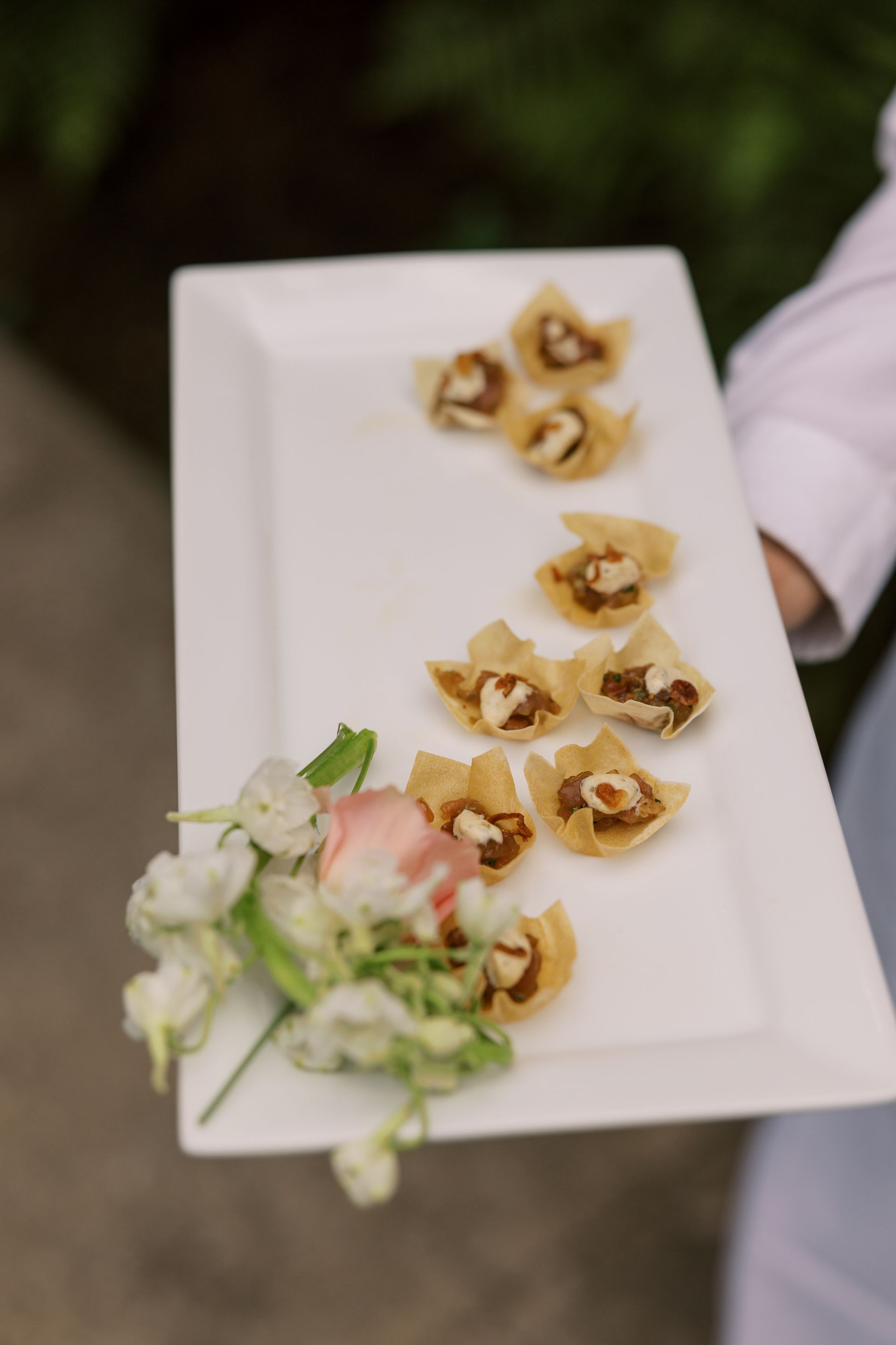 A server holding a white platter with appetizers and a small floral arrangement. The appetizers are golden-brown, cup-shaped, and filled.