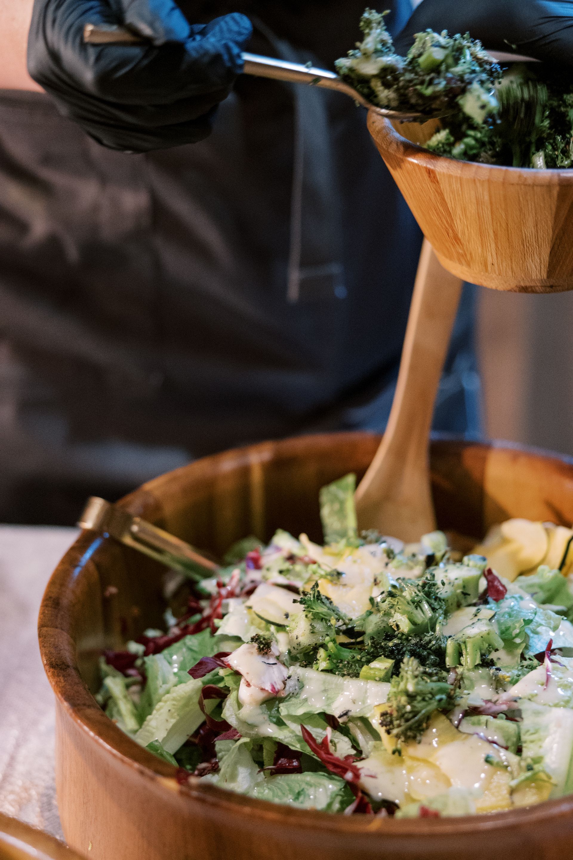 Person in black gloves adding salad to a large wooden bowl. Green and red lettuce, and dressing are visible.