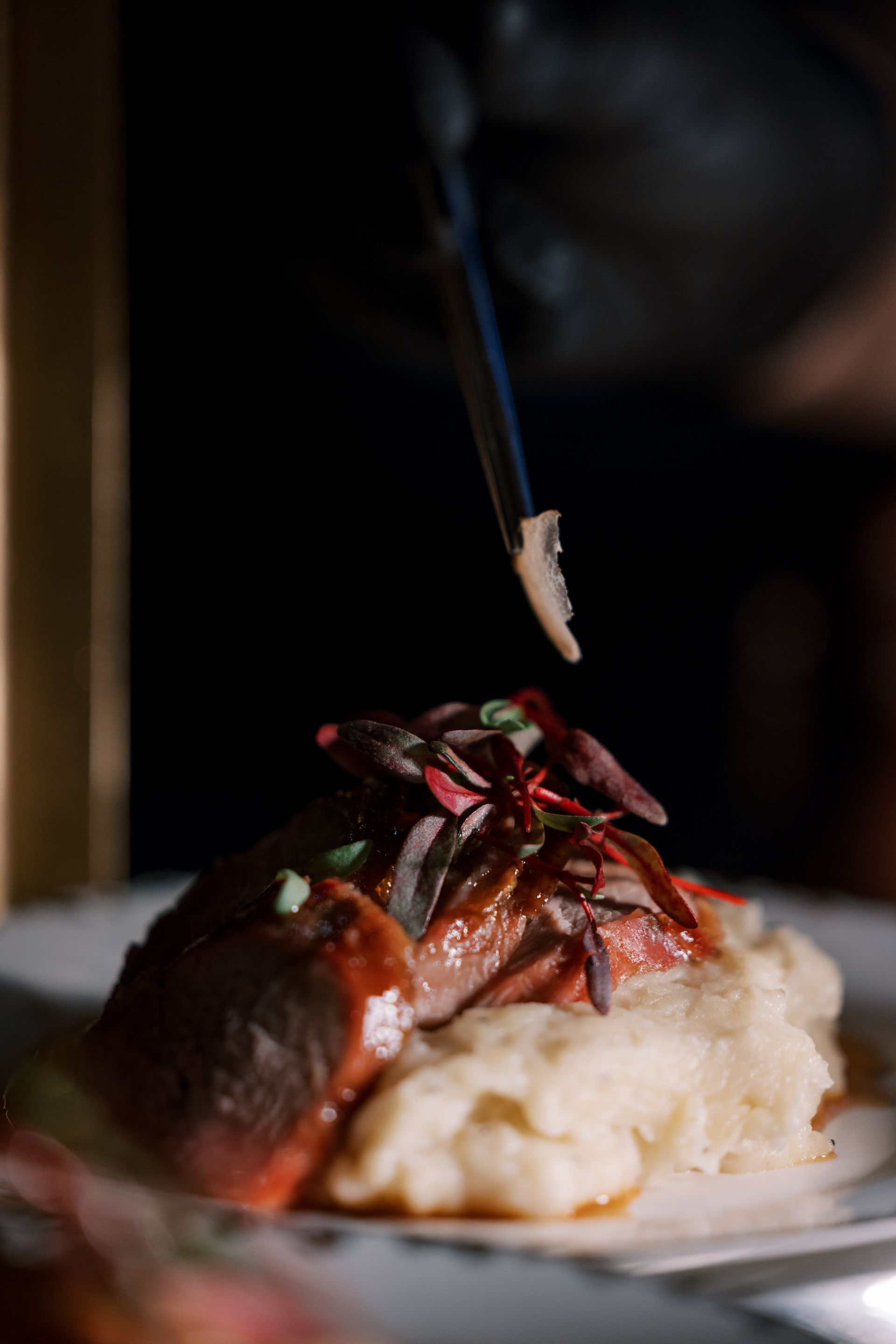 A plate of food, likely a meat dish with mashed potatoes and garnish. Tongs are about to place a final piece of food.