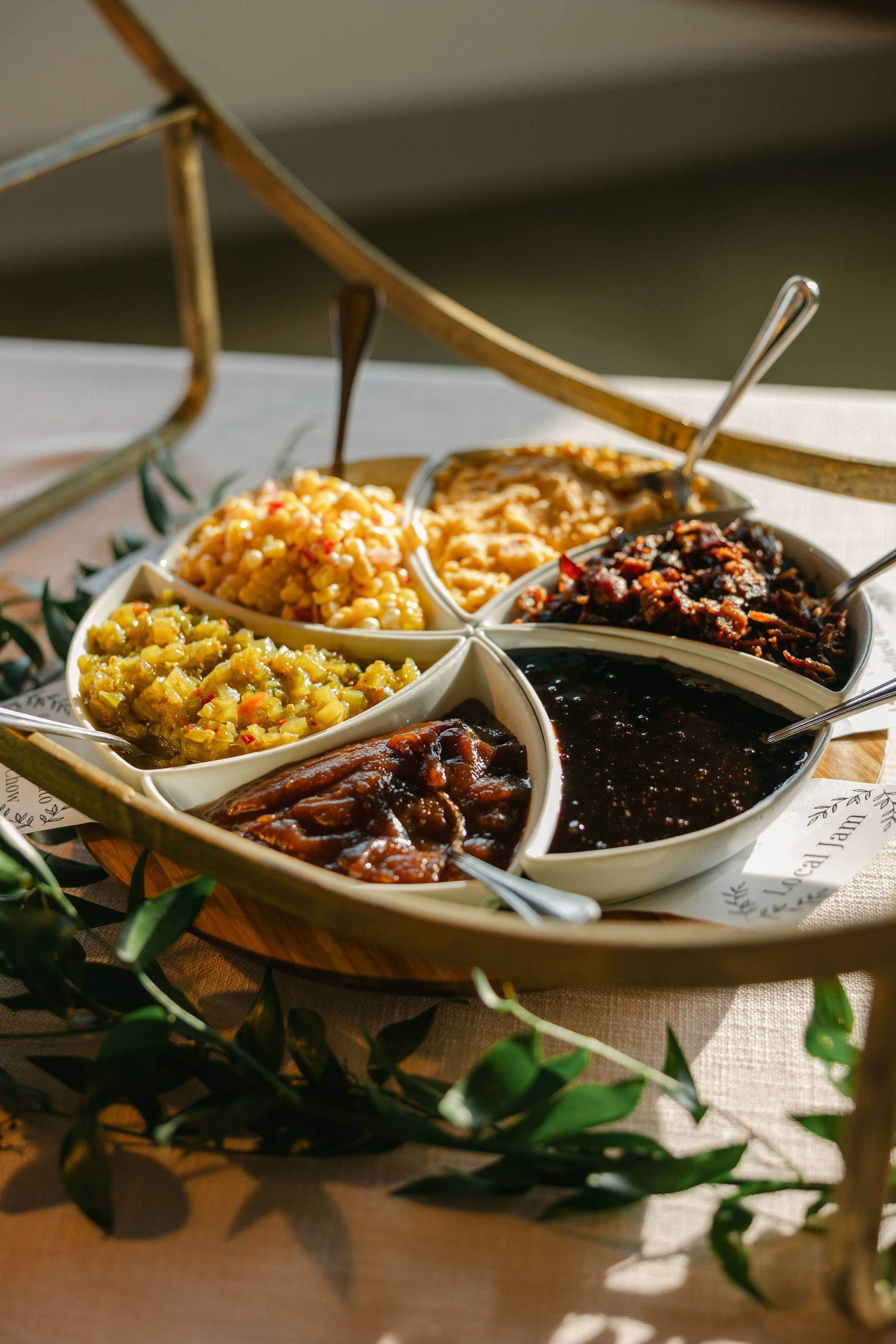 A tiered serving tray holding bowls of different colored chutneys and sauces, set on a table decorated with greenery.