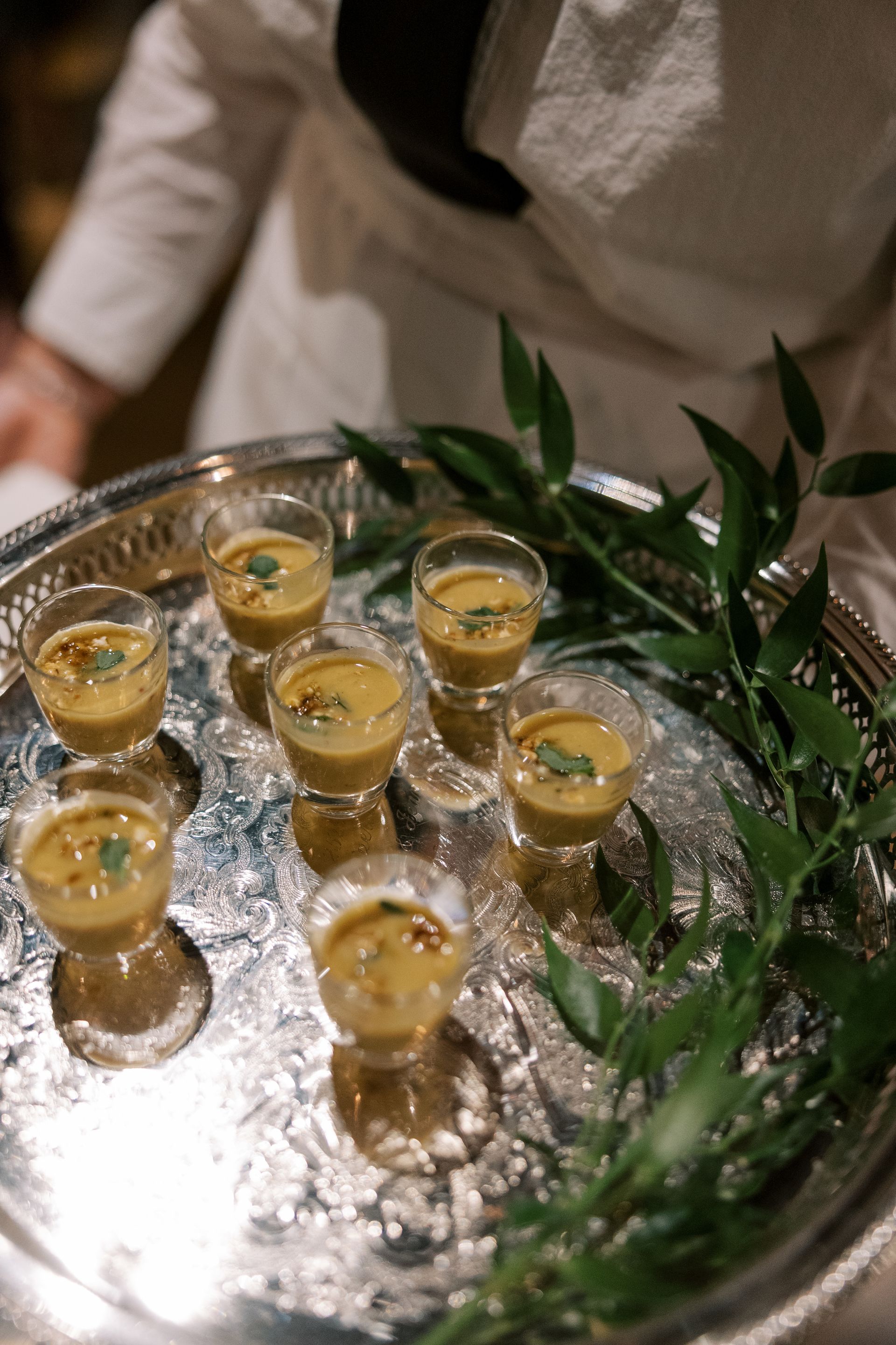 A server holding a silver tray of small soup shots, garnished with herbs, set against a white uniform.