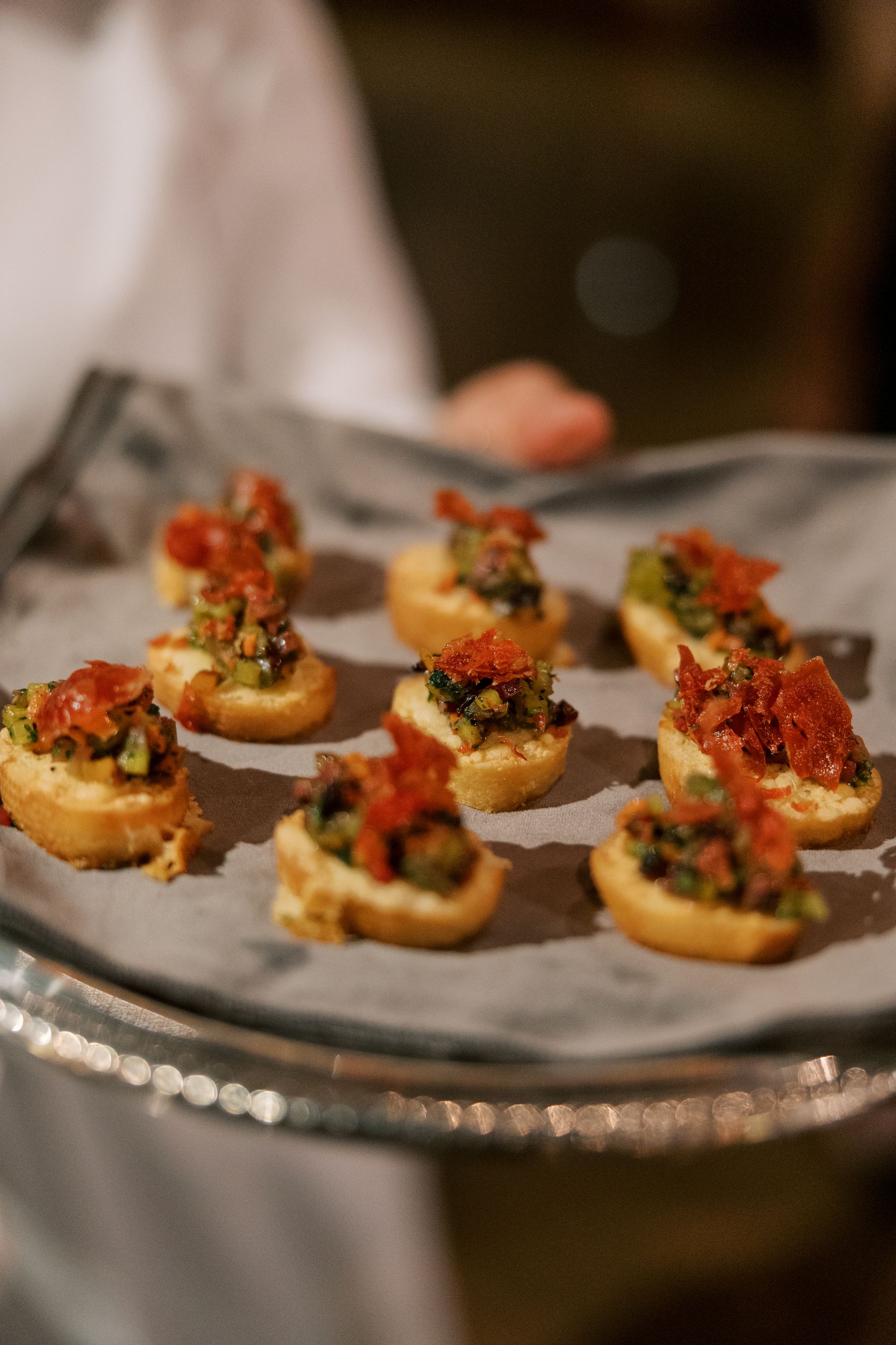 A server holds a tray of bite-sized appetizers: toasted bread topped with green olive tapenade and red sun-dried tomatoes.