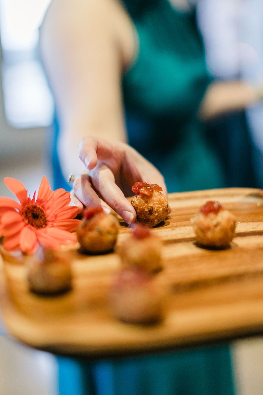 A person in a teal dress offers a wooden tray of fried food topped with red relish, alongside an orange flower.