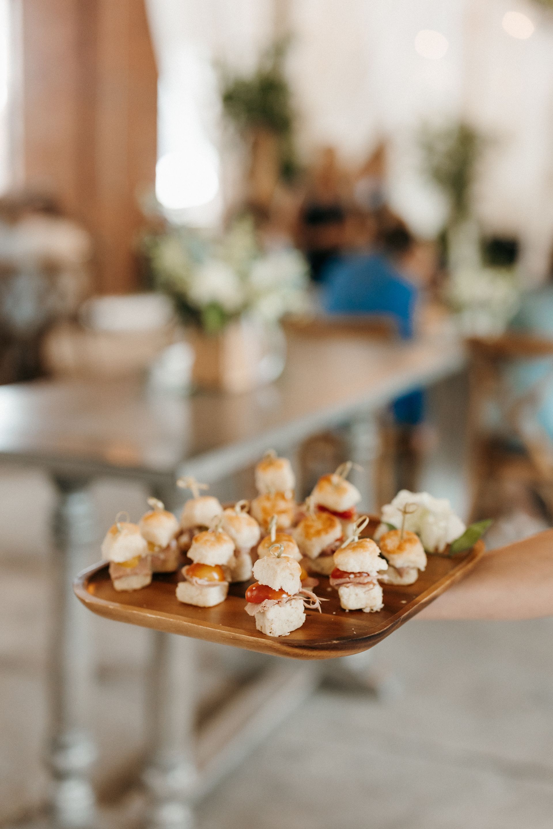 A hand holding a wooden tray with mini sandwiches topped with toothpicks. Blurred background of a room with tables.