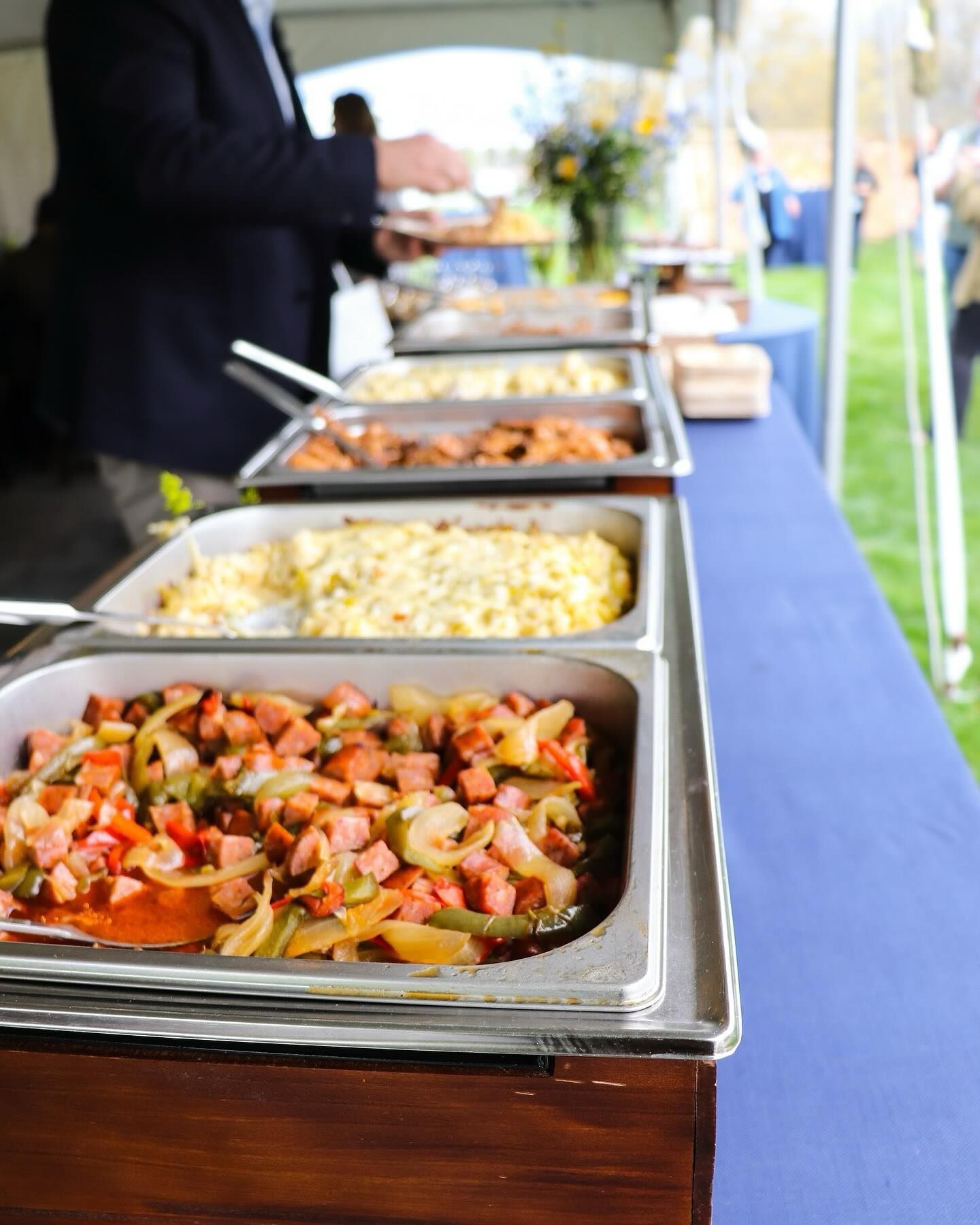 A buffet table laden with various food dishes, including a sausage and vegetable mix, attended by a person in a blazer.