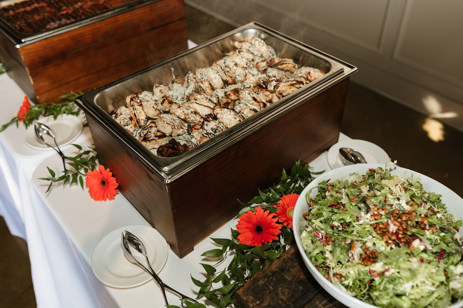 A buffet table with trays of food, including grilled chicken, and a large salad. Decorated with flowers and greenery.