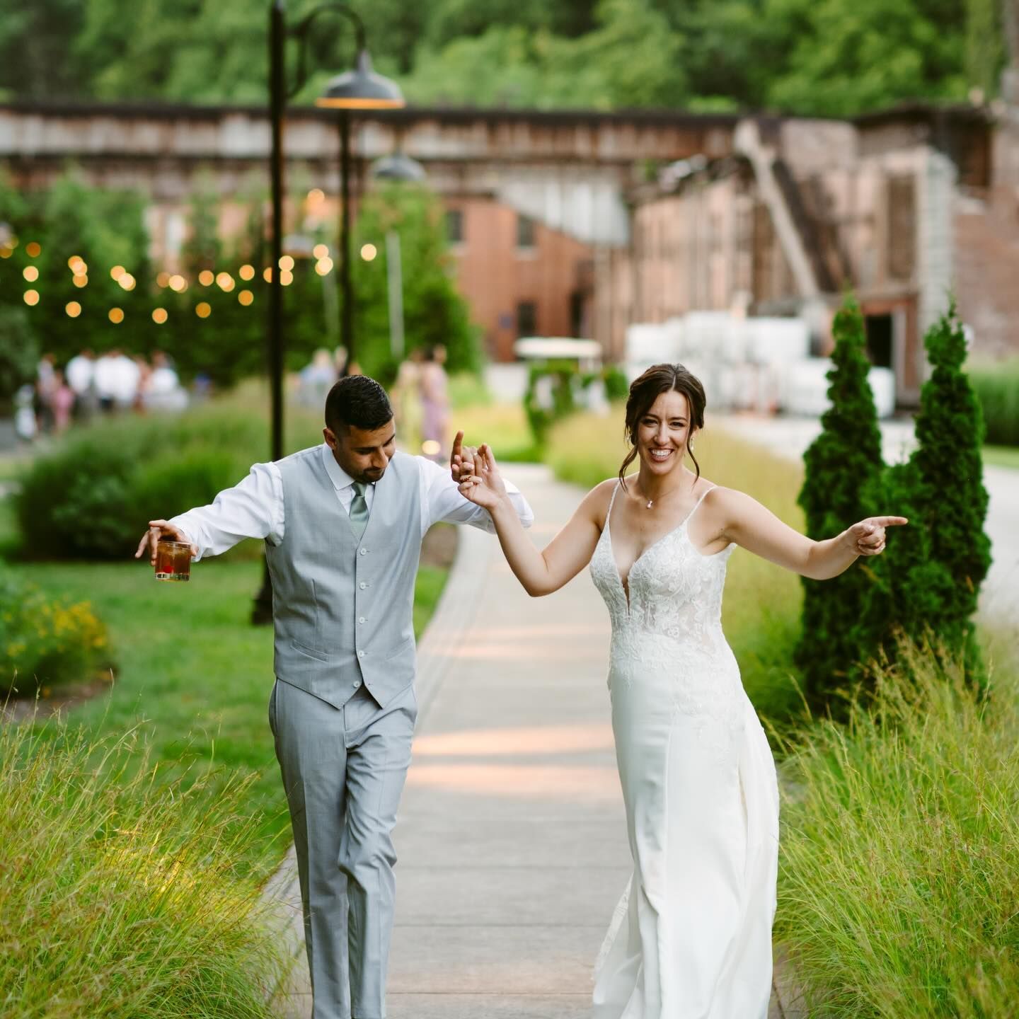 Newlyweds joyfully walk, holding hands. The woman points, the man holds a drink. Outdoor wedding setting.