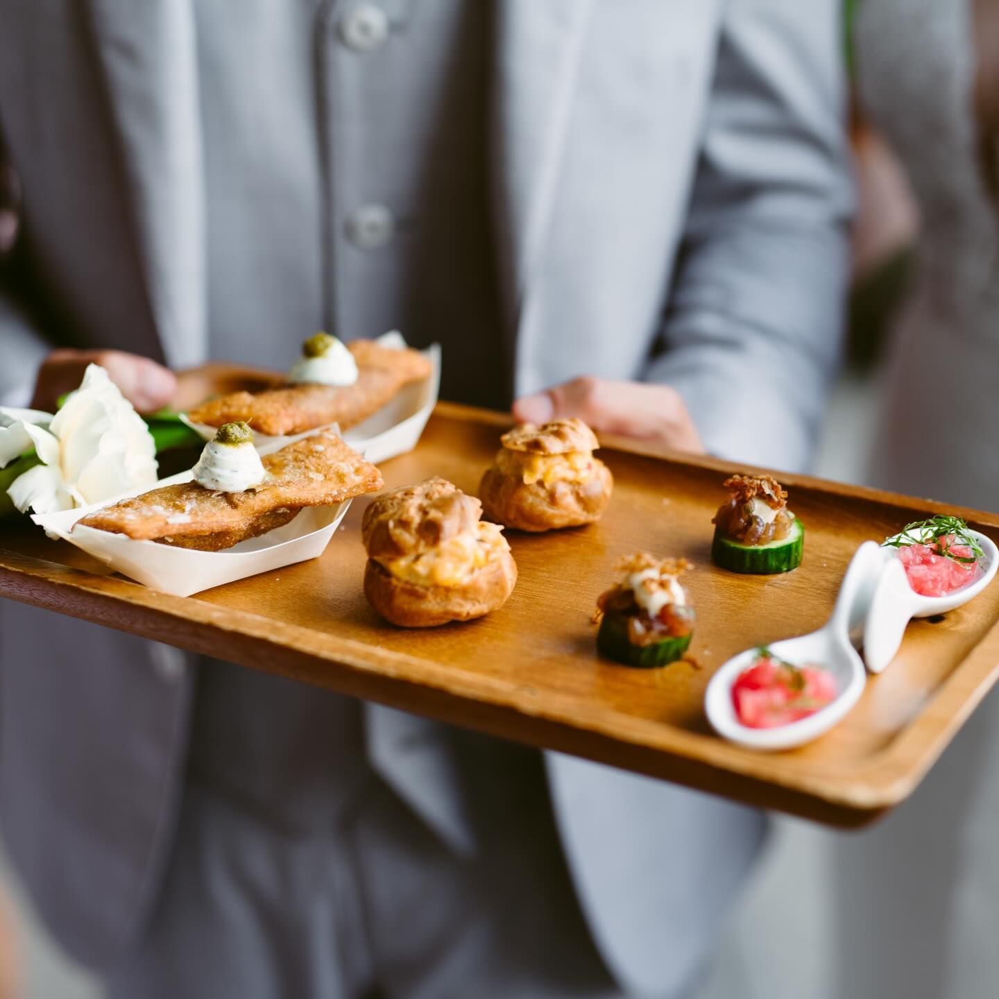Person in gray suit holding a wooden tray with appetizers: mini pastries, fish bites, and cucumber rounds.