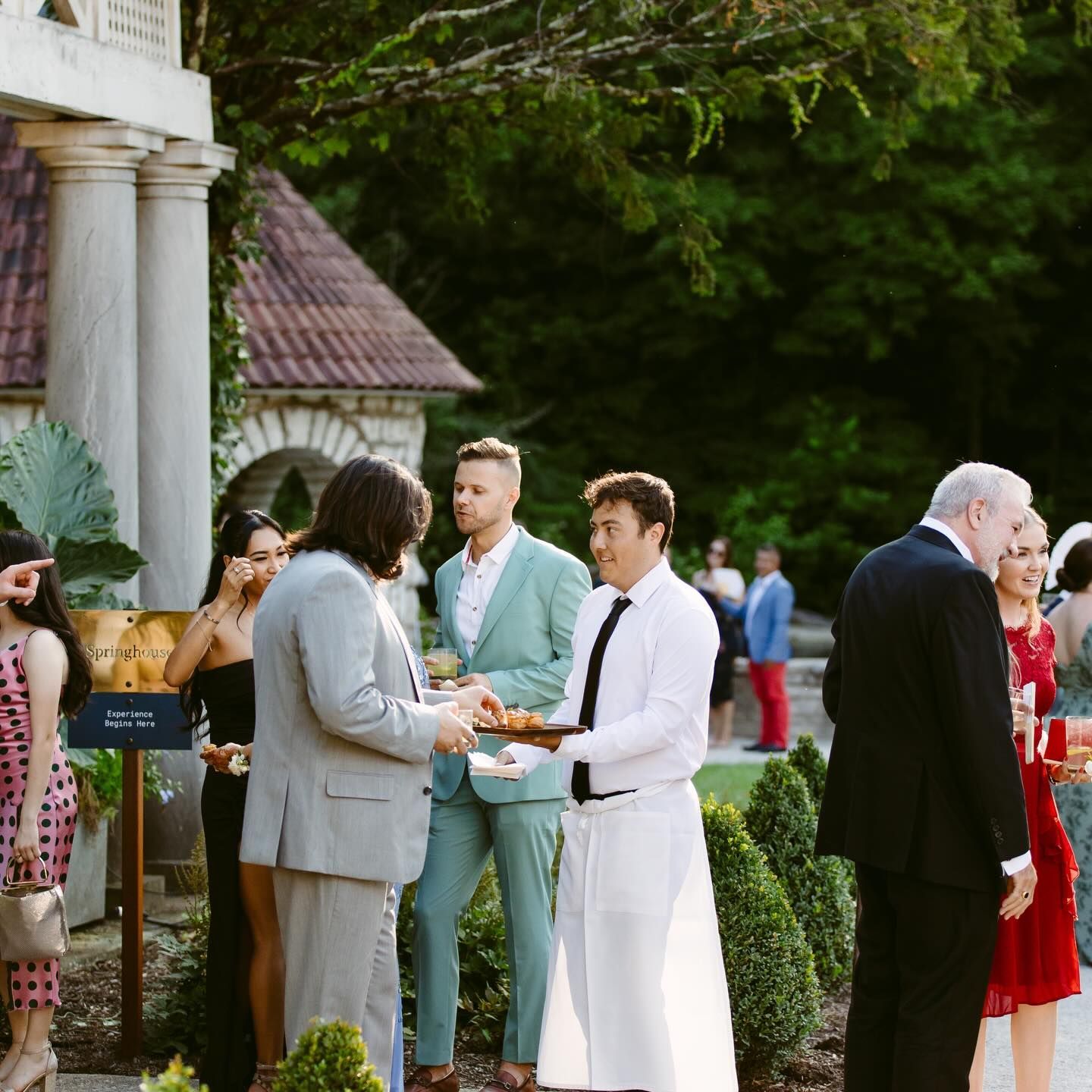 People at outdoor event. A server offers appetizers. Guests in formal attire mingle on a lawn.