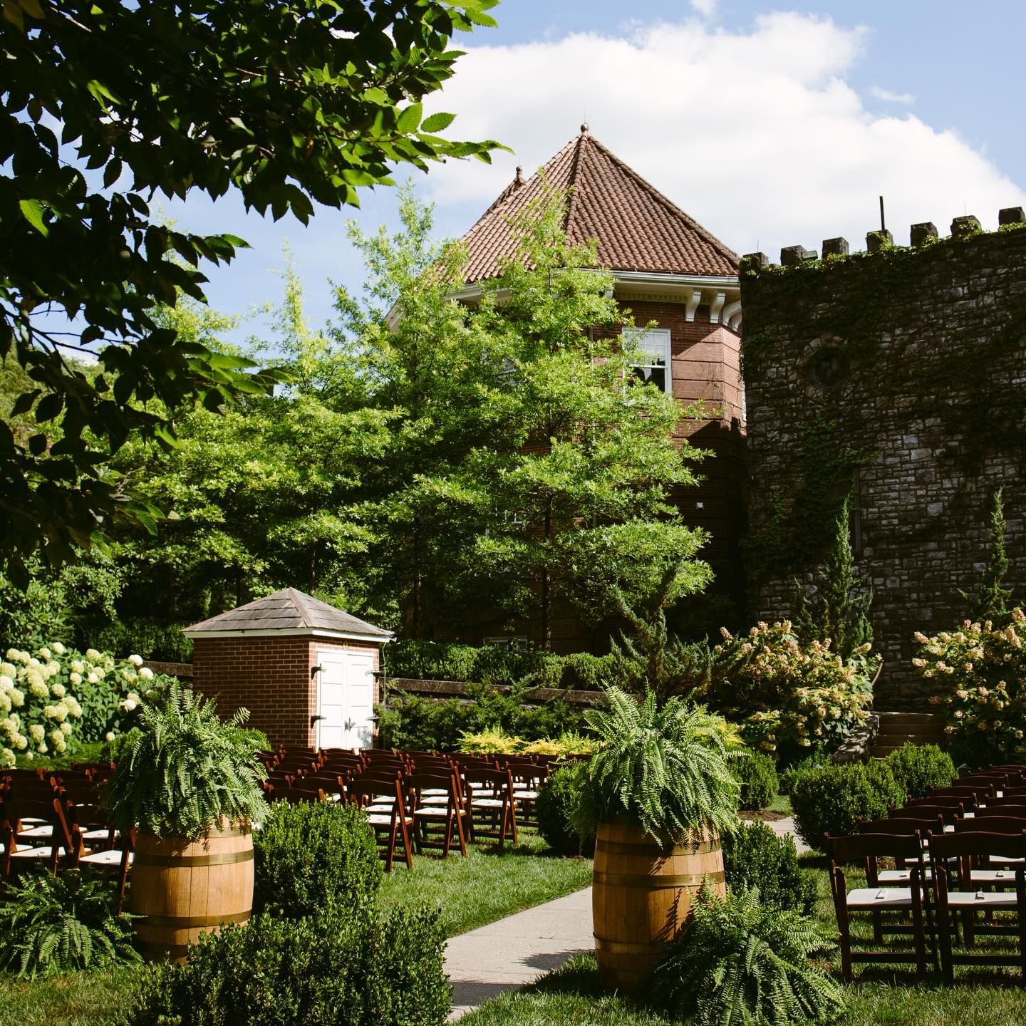 An outdoor wedding ceremony setting, with wooden chairs, greenery, and a brick building backdrop.