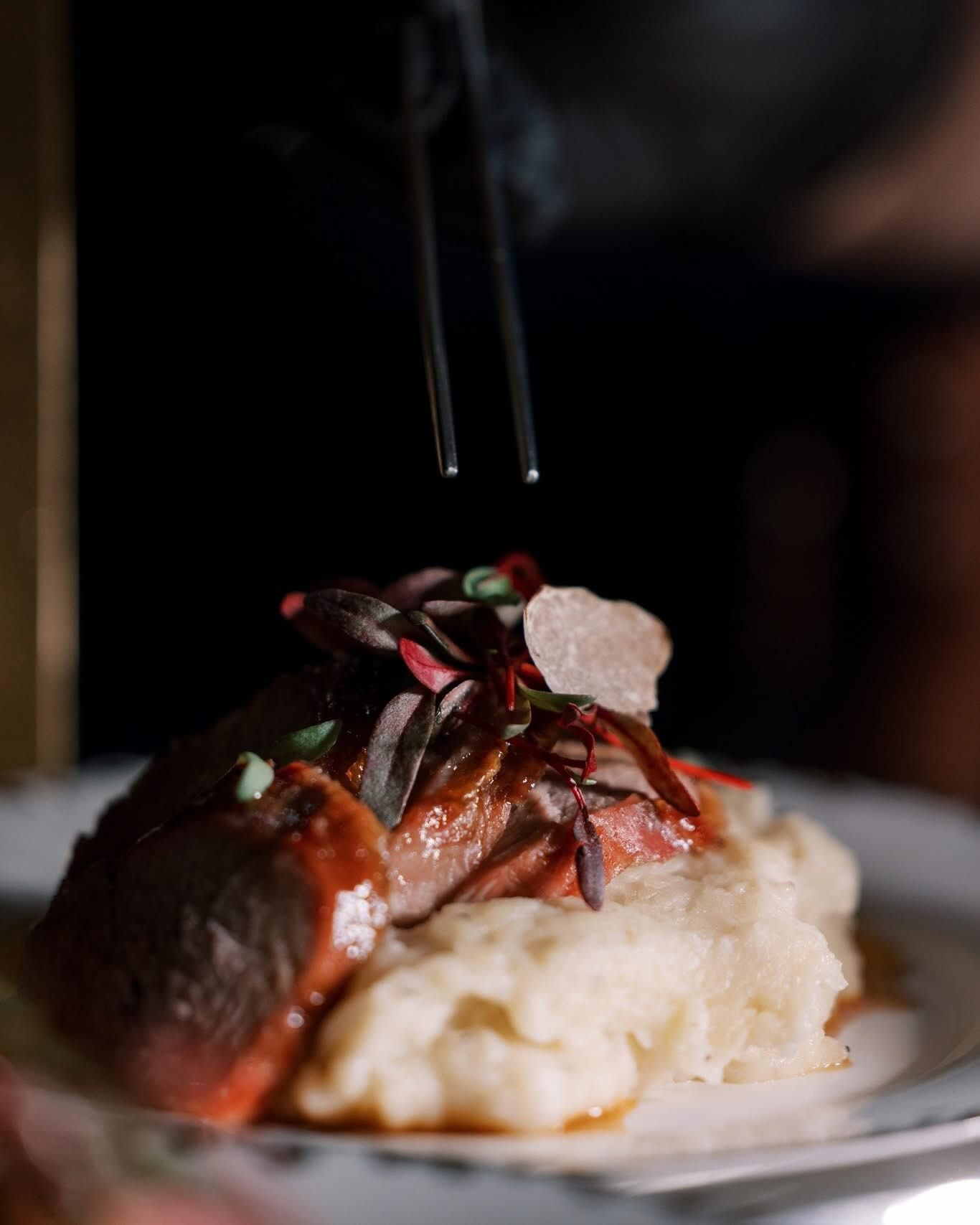 Seared meat atop mashed potatoes, garnished with microgreens. Smoke rises from tongs, likely for presentation.