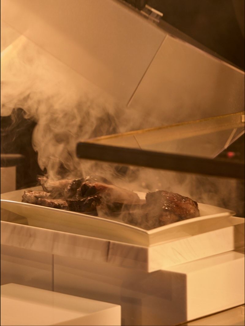 Steaming cooked meat on a white rectangular plate, under a warming light.