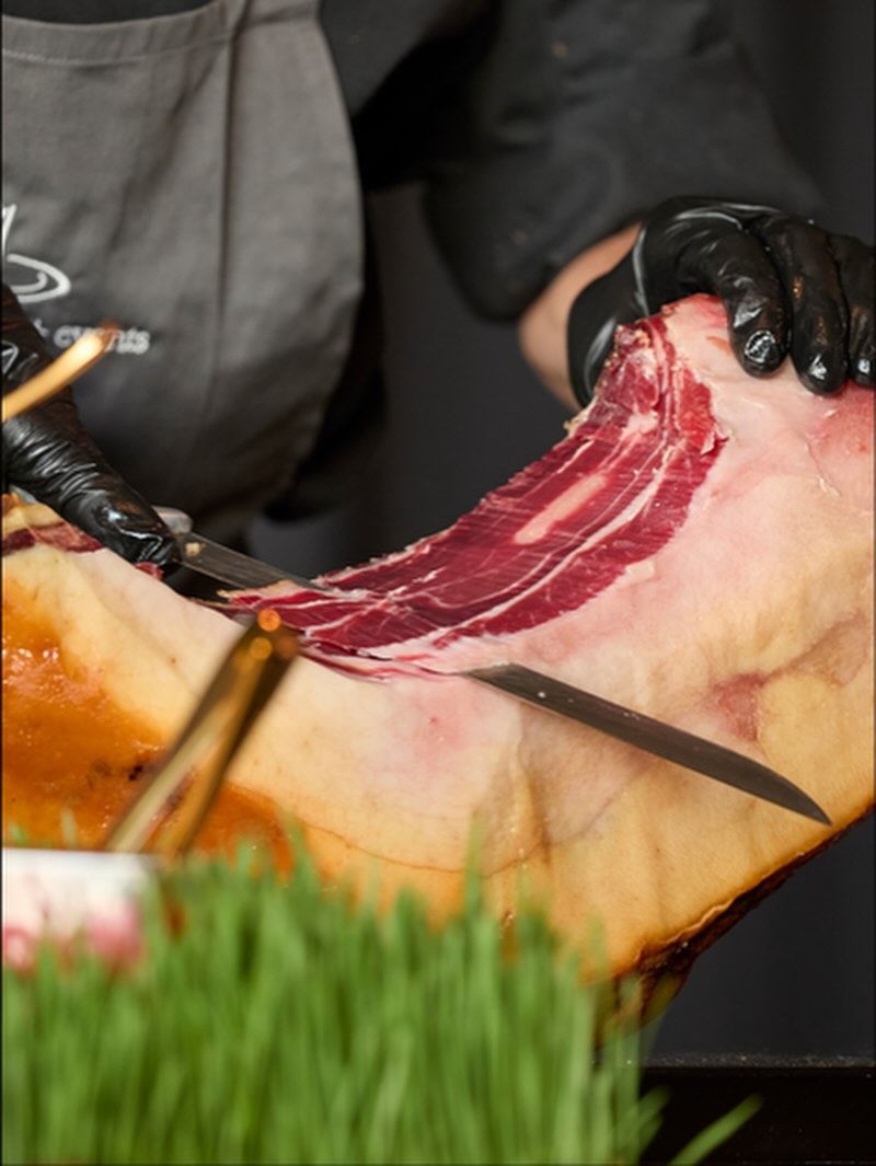 Person wearing black gloves slicing cured meat with a long knife.