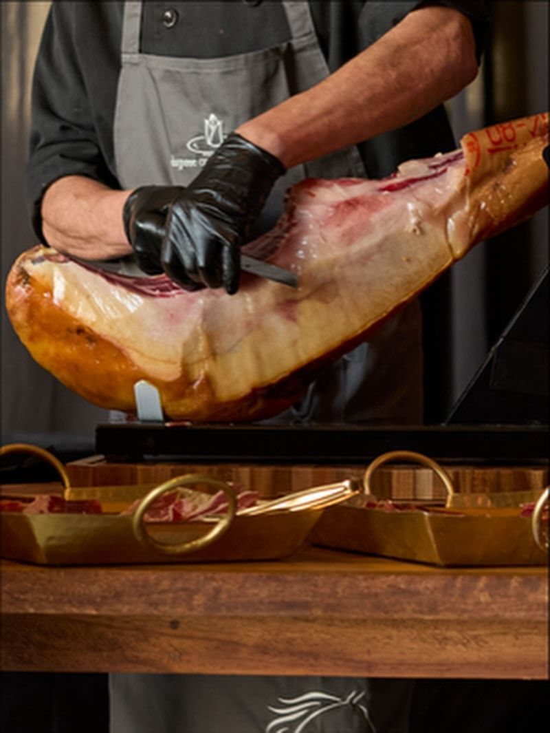 A person in black gloves carving jamón with a knife, on a wooden surface with gold trays.