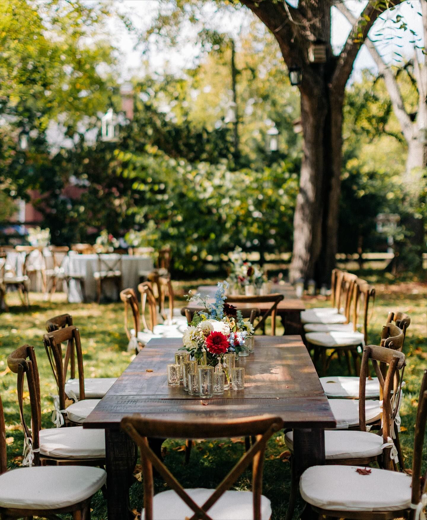 Outdoor wedding reception with wooden tables set for guests; floral centerpieces.