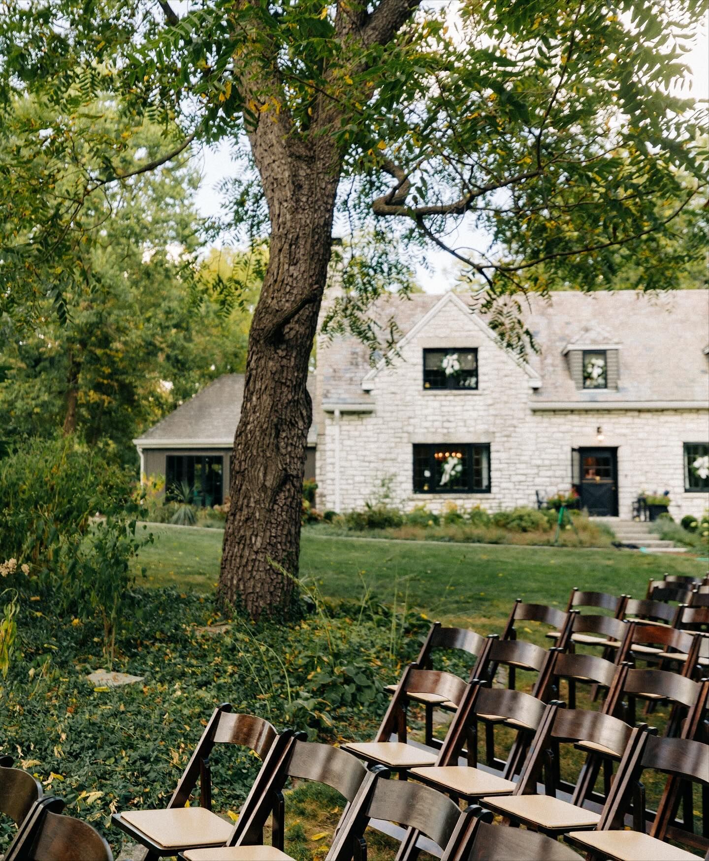 Brown chairs face a stone house with trees overhead, prepared for an outdoor event.