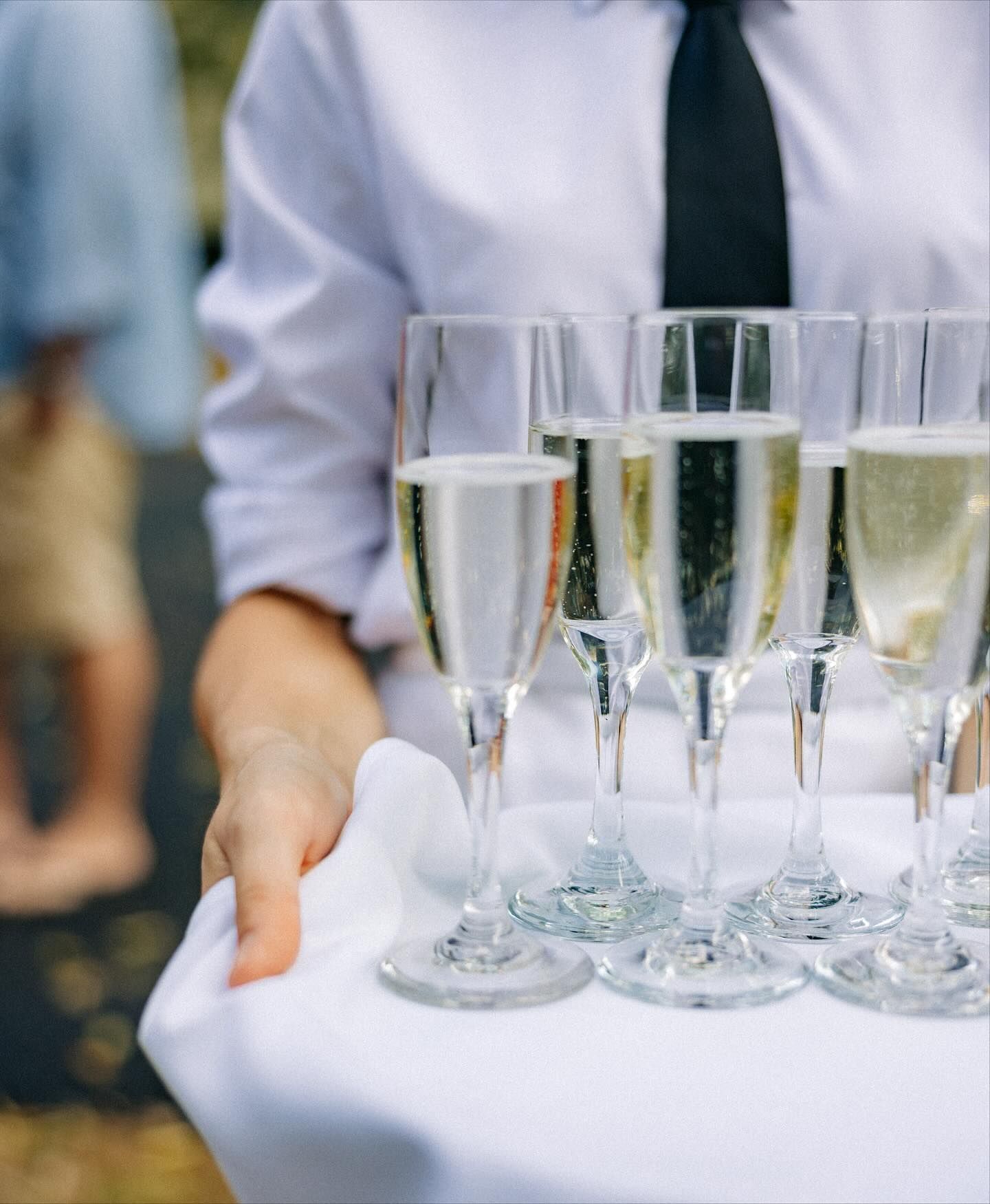 Server holding a tray of champagne flutes on a white cloth; person in white shirt and black tie in the background.
