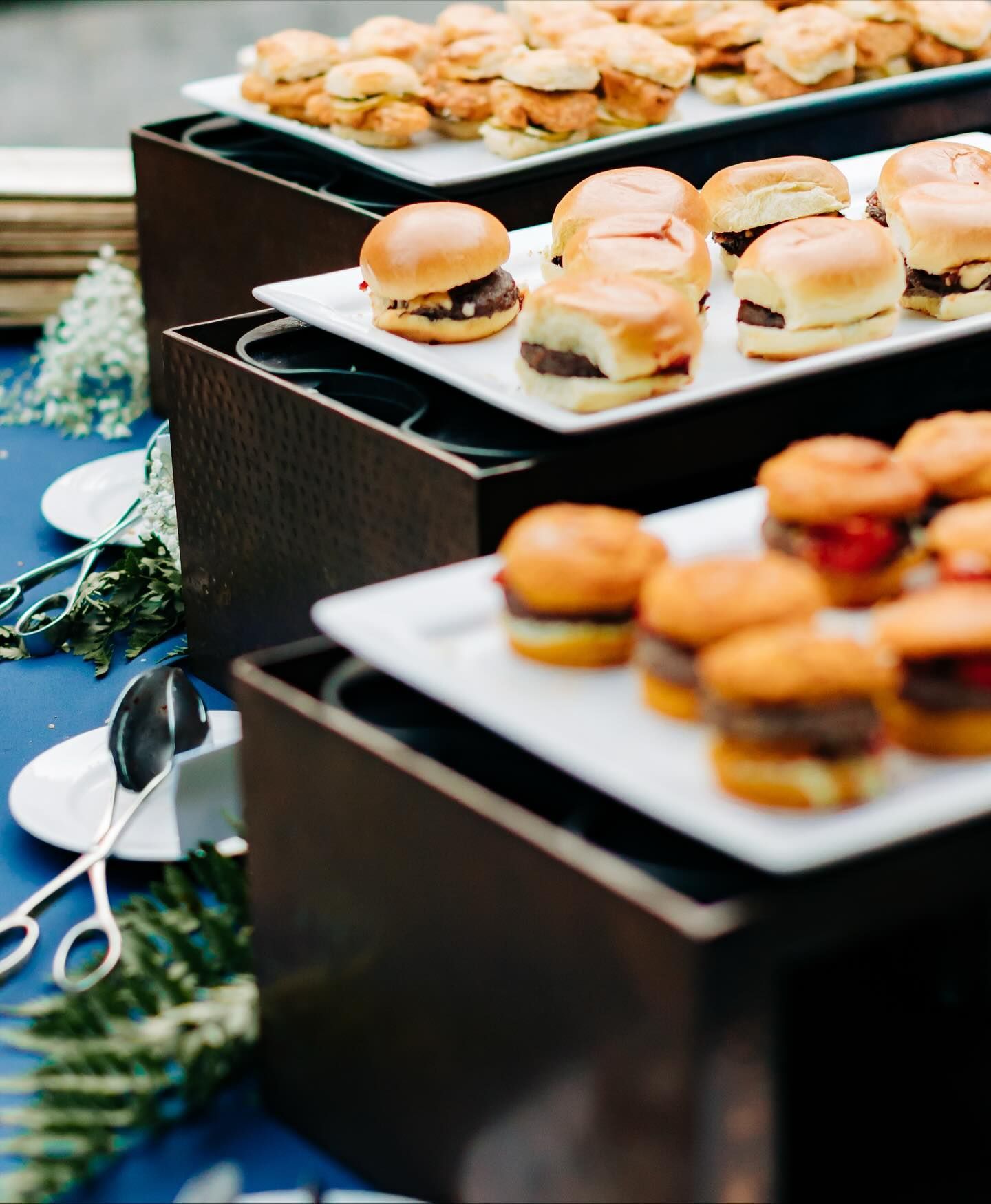 Buffet with sliders displayed on tiered serving trays. Blue tablecloth with silver utensils and greenery.