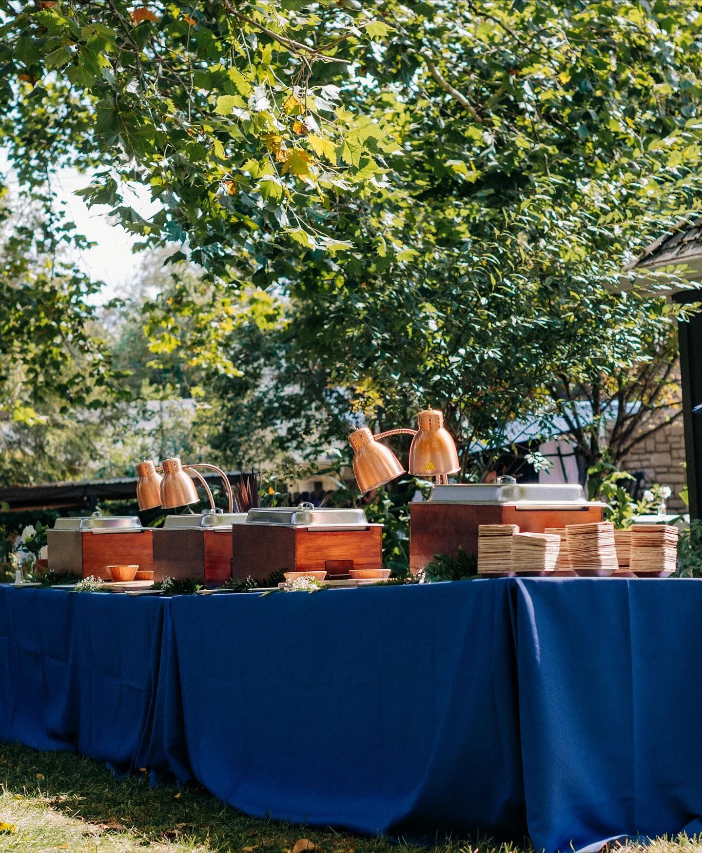 Buffet table with navy tablecloth, serving food outdoors under a leafy tree.