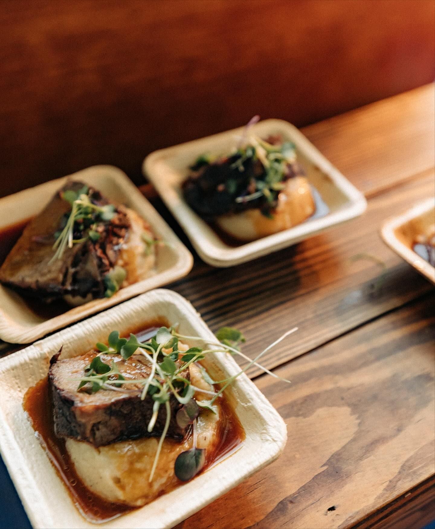Small dishes of food on a wooden surface, each with meat, mashed potato, and sprouts.