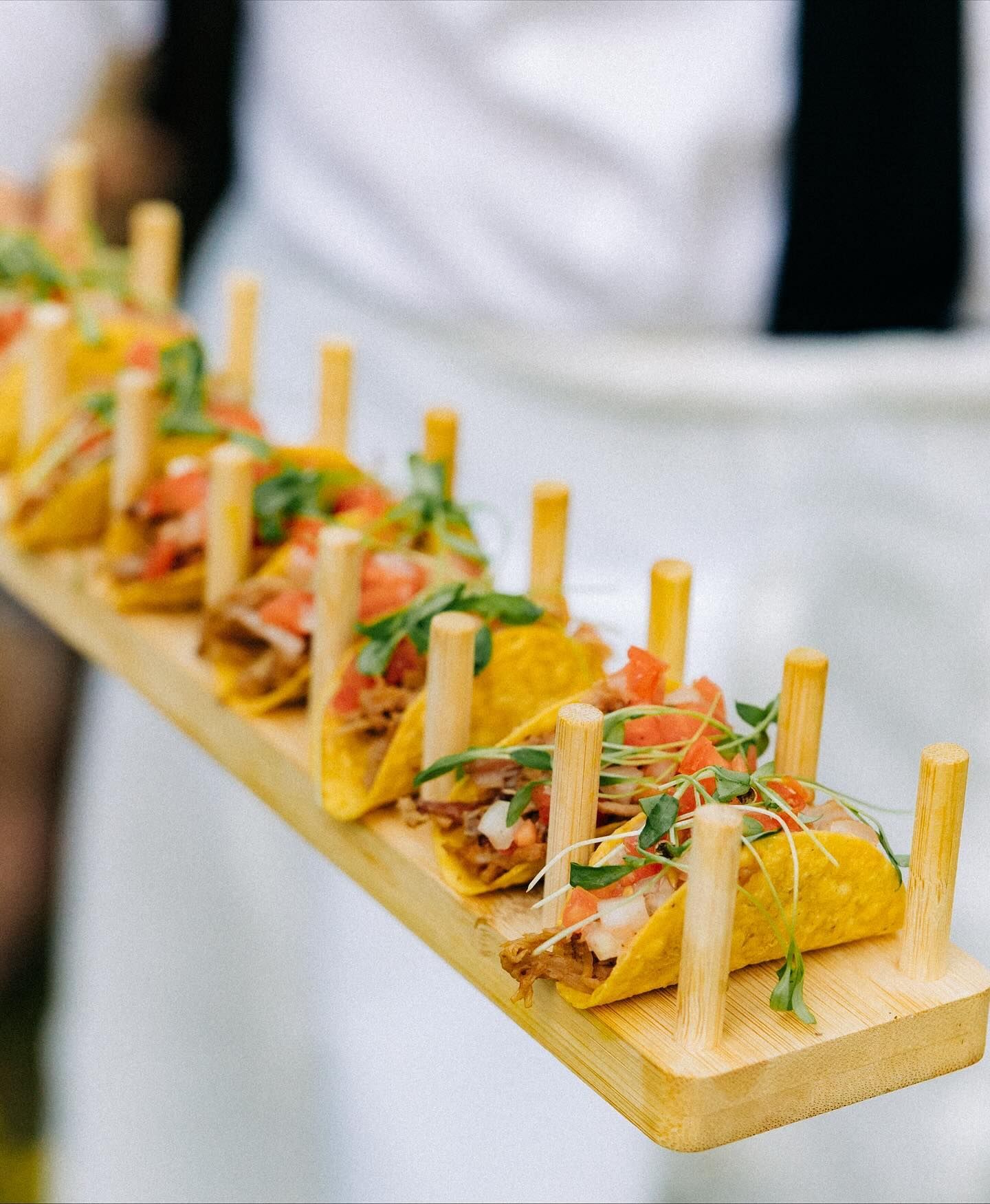 Wooden tray with a line of tacos, each secured by a wooden stick.
